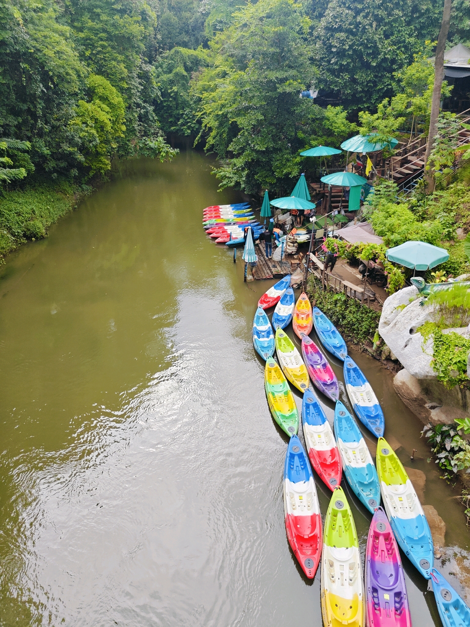An aerial view captures a river flowing through a lush, green forest. Along the riverbank, a collection of brightly colored kayaks are lined up. Some kayaks are red, blue, yellow, green, and purple, while others are a mix of these colors. A wooden dock extends into the water, where several people are visible, some standing and others seated. Several green umbrellas are set up near the dock, providing shade. The water is murky brown, reflecting the surrounding foliage. The riverbanks are densely covered with trees and vegetation, creating a natural, serene atmosphere. On the right side of the image, large white rocks are covered with moss and greenery. A decorative lizard statue is perched on one of the rocks. The scene appears to be a recreational area, likely a place for kayaking and outdoor activities, located in Mu Si, Thailand.
