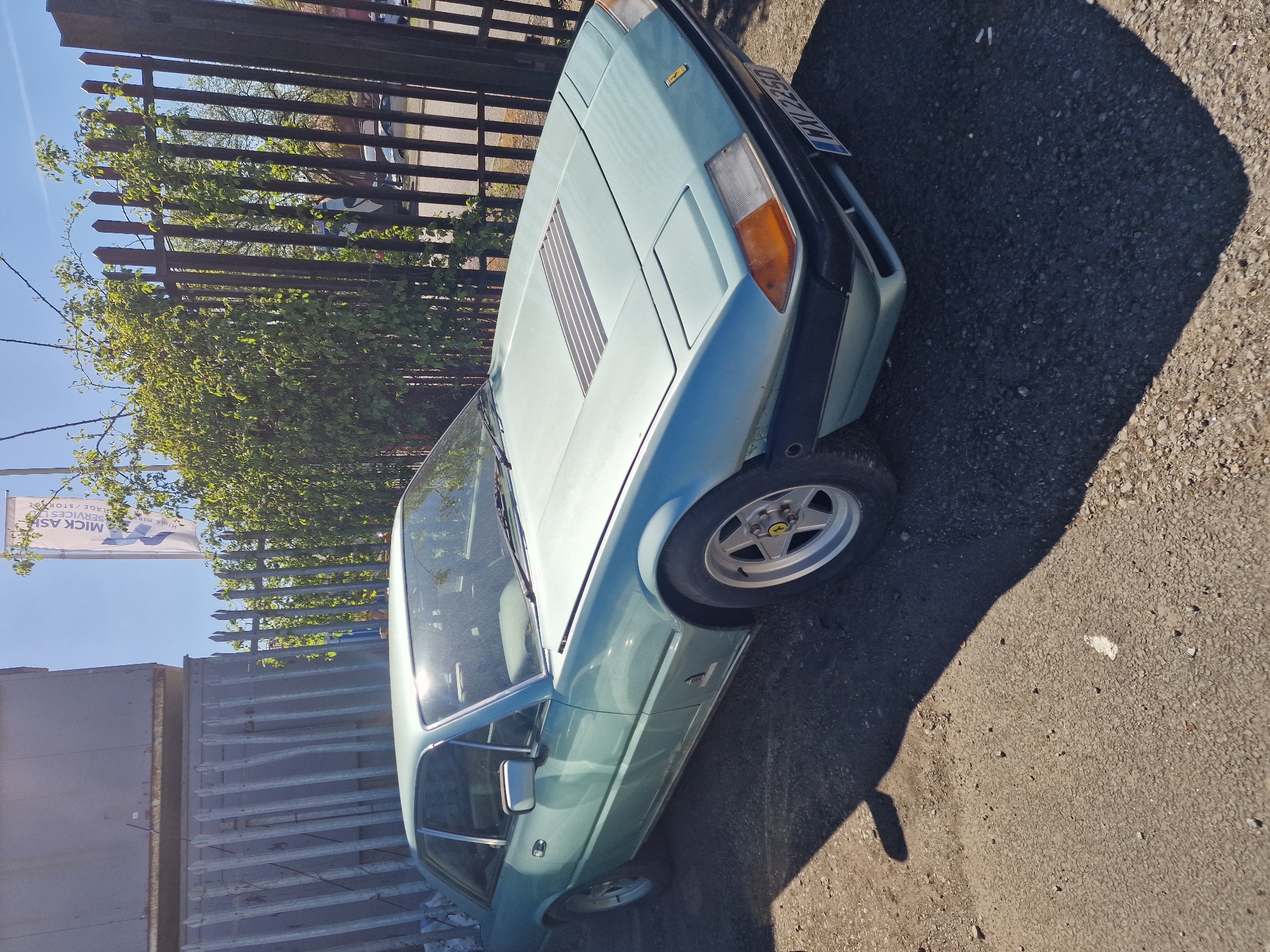 A light blue vintage Ferrari, likely a 400 or 412 model, is parked outdoors on an asphalt surface under clear, sunny daylight. The car is viewed from the front-right quarter, showing its angular design, pop-up headlights (retracted), and a rectangular louvered vent on the hood. The front bumper is dark, and the front right wheel, with a silver five-spoke design and a visible Ferrari prancing horse logo on the hubcap, is prominently featured.
Strong shadows are cast on the ground to the right of the car, indicating a high sun position. In the background, a dark brown metal fence runs horizontally, behind which dense green foliage of bushes or trees is visible. Further in the background, a light-colored commercial banner is partly visible against the blue sky, displaying the text "MICK ASH" and partially "SERVICES / STORAGE". Another segment of the banner reads "HIRING".
The vehicle bears a UK registration plate on its front, which appears to read "D 923 MXY", indicating a registration from late 1986. The setting suggests a commercial or light industrial area, consistent with the fence and business signage, within South Tyneside, United Kingdom. The image captures a static scene with no discernible activity or emotions.