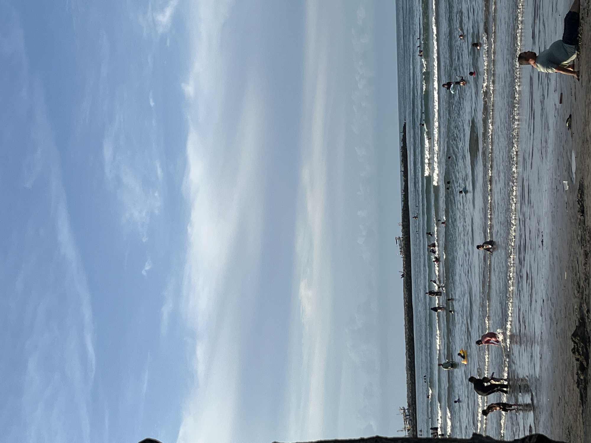 The image shows a beach scene in Lhokseumawe, Indonesia. A crowded beach with people swimming and wading in the ocean is depicted. Waves are visible, and a concrete barrier or pier extends into the water. The sky is blue with some scattered clouds. One man is crouched near the edge of the beach.