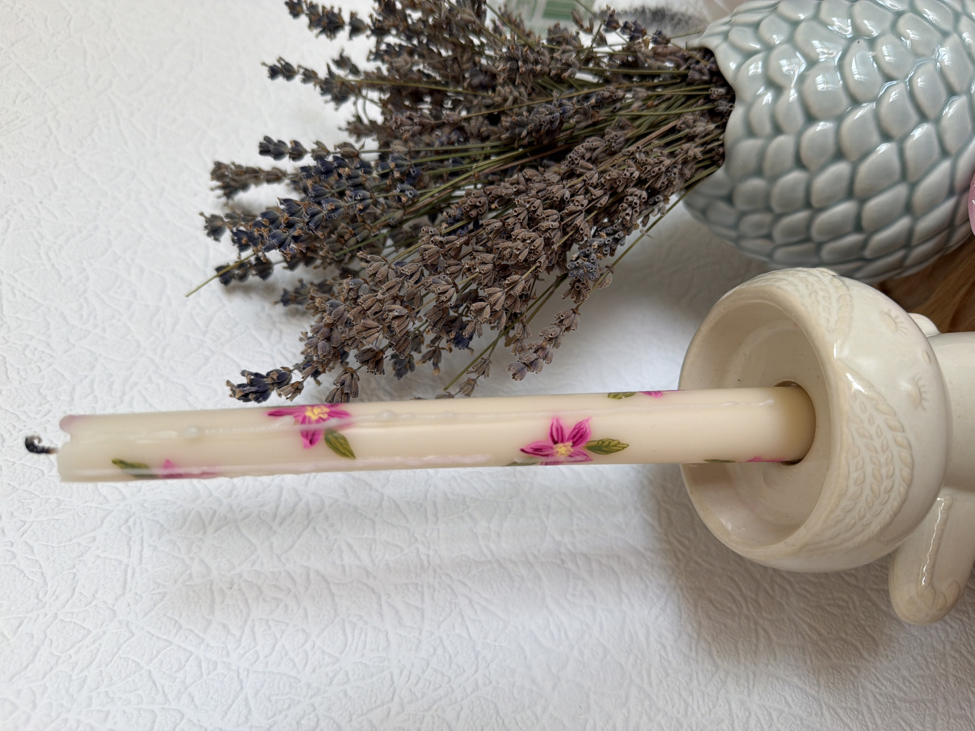 The image depicts a close-up still life of decorative items. A slender, cream-colored candle with painted pink flowers and green leaves is placed in a light-colored ceramic candleholder, which has embossed detailing resembling feathers and closed eyes. To the right of the candle, a light blue, textured vase, shaped like a pineapple, holds a bunch of dried lavender. The background is a textured, white wall. The scene is lit by natural light, creating soft shadows. There is no indication of a specific location in Kyiv, Ukraine.