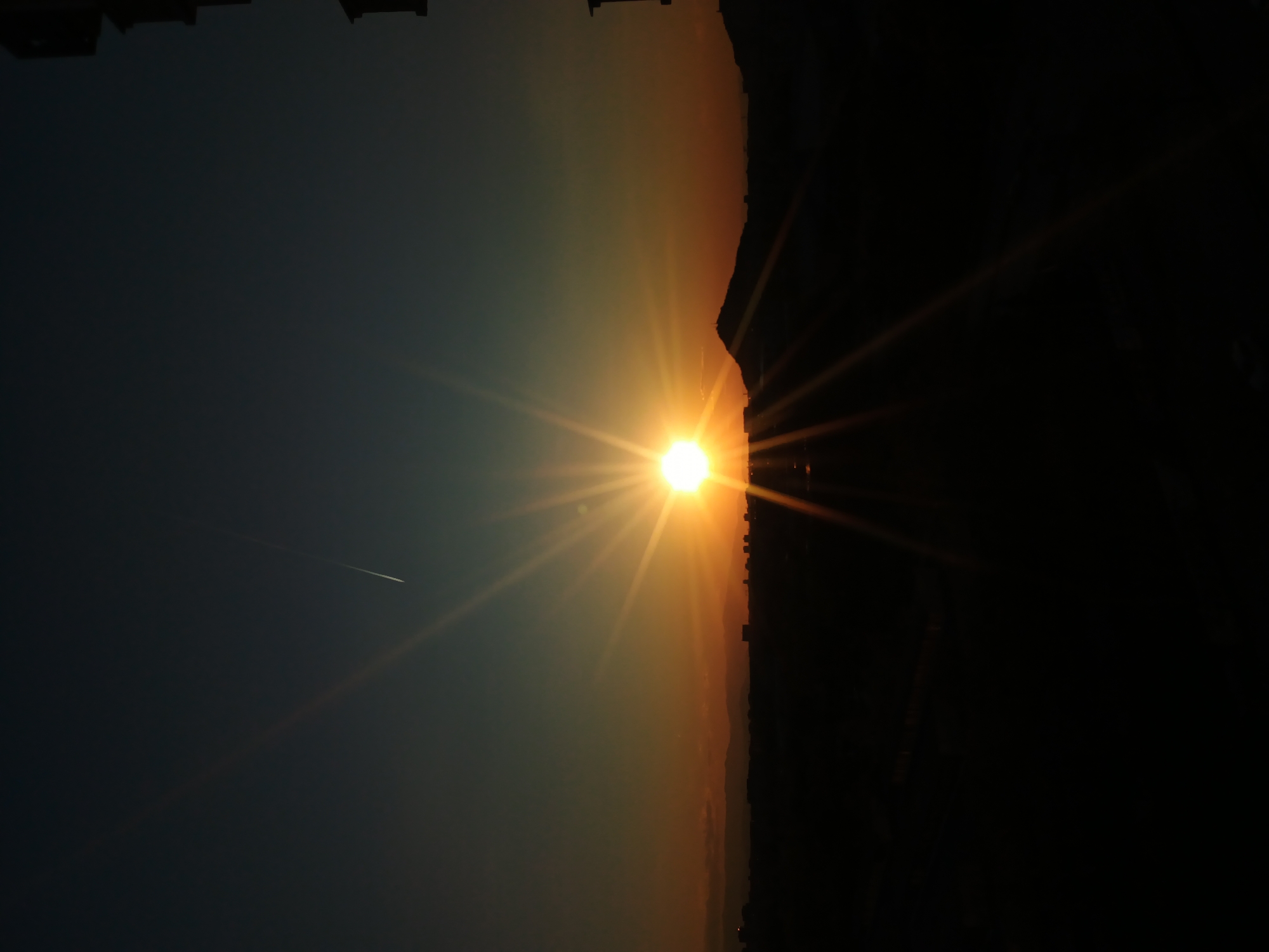 The image shows a bright sun setting on the horizon over a city, likely taken in Plovdiv, Bulgaria. The sun's glare is intense, radiating beams of light across the sky. To the left, the sky transitions from a deep blue to a lighter gradient near the horizon. A faint contrail from an airplane is visible against the blue sky. To the right, silhouetted buildings and potentially hills are present, with the rightmost area being completely dark. The horizon line where the sun sets appears to be on or near the city's edge.