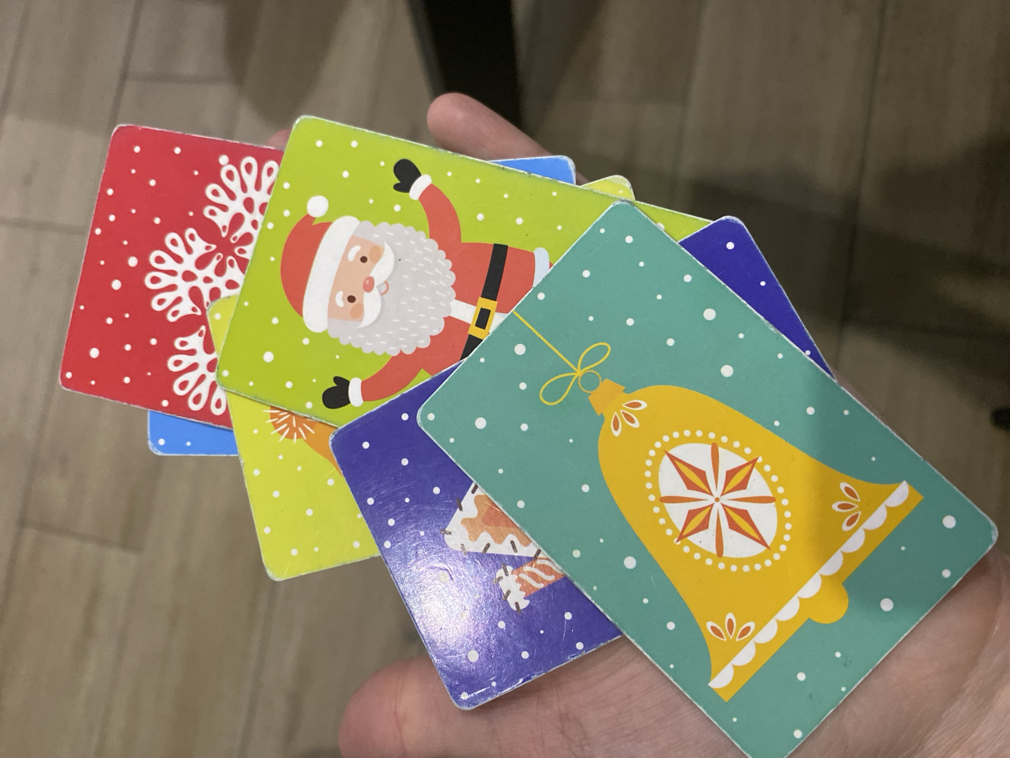 The image shows a hand holding five square cards with Christmas-themed illustrations. The cards feature a red background with white snowflakes, Santa Claus on a light green background, a light green background with a gold Christmas bell, a purple background with a candy cane illustration, and a light green background with white dots. The cards are slightly overlapping and appear to be used. The background is blurred and shows a light brown surface. The city of Chaikovsky, Russia is not apparent in this image.