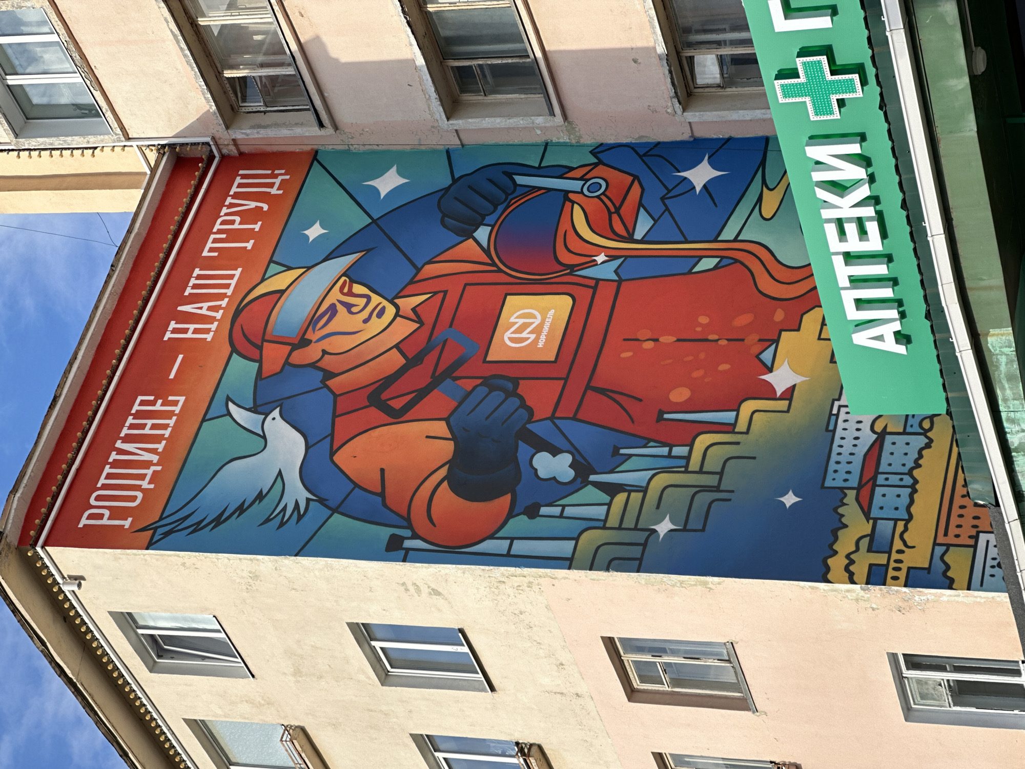 This media file depicts a mural painted on the side of a building in Monchegorsk, Russia. The mural is a vibrant, stylized representation of a worker in a red and orange suit, wearing a helmet and blue gloves. The worker is holding a tool, possibly for industrial work, and is depicted against a backdrop of blue and yellow abstract shapes, stars, and a cityscape. A white dove is also present in the mural, a symbol often associated with peace. The overall impression is one of strength, labor, and perhaps a nod to industrial heritage.
Above the mural, on the building's facade, there is text in white Cyrillic letters on a red background that reads "РОДИНЕ – НАШ ТРУД!" which translates to "To the Motherland – Our Labor!". To the right of the mural, a green sign is visible with white Cyrillic text that reads "АПТЕКИ", meaning "Pharmacies", and a green cross symbol, indicating the presence of a pharmacy.
The image appears to have been taken during the daytime, with clear blue skies visible. The lighting suggests it is a sunny day. The building itself is an apartment building with several windows. The perspective is from below, looking up at the mural and the building. The style of the mural suggests a mid-20th century Soviet aesthetic, emphasizing collective labor and patriotism.