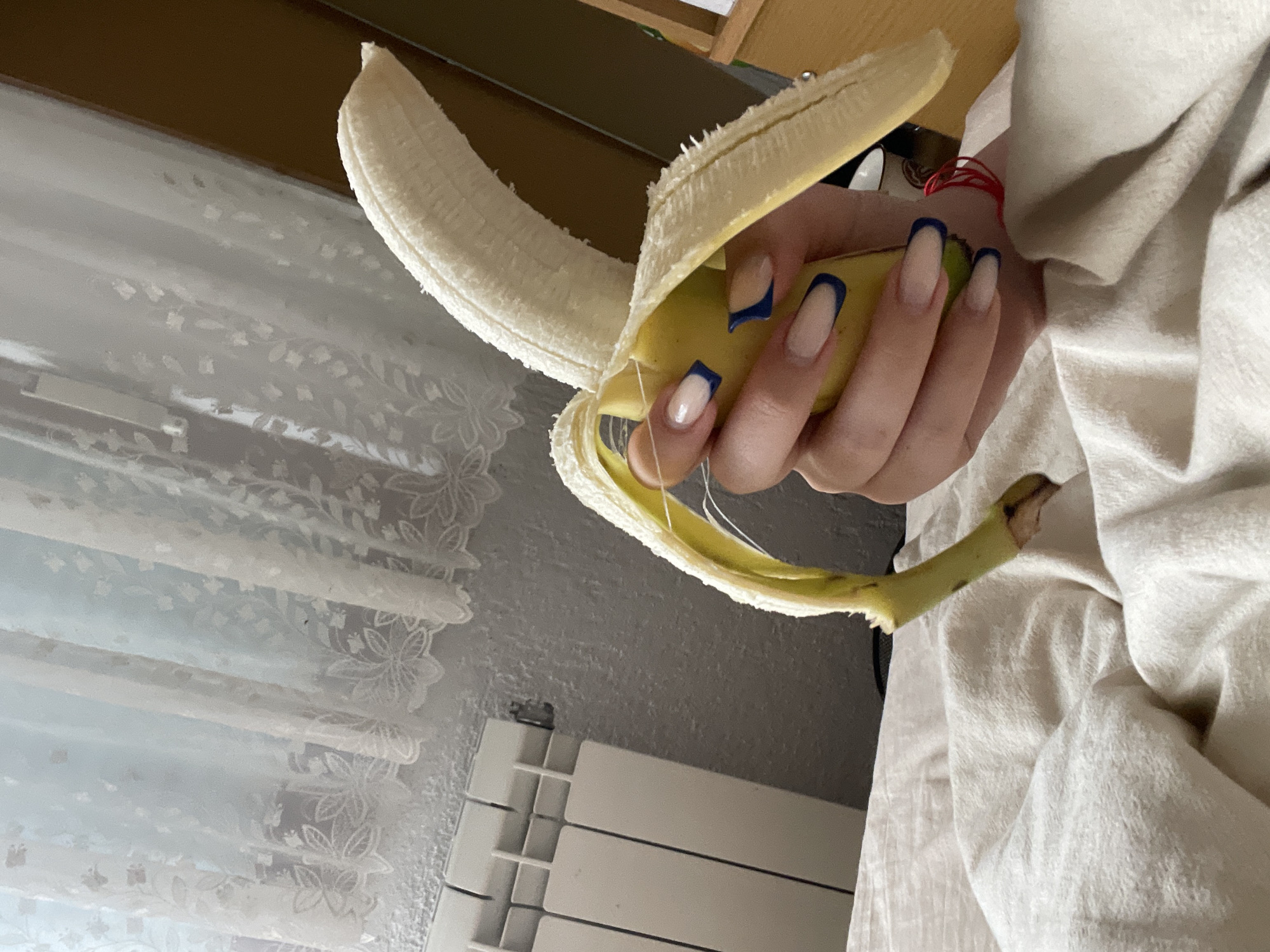 A partially peeled banana is held in a hand with long nails painted with a blue french manicure. The background includes a window with lace curtains, a radiator, and a section of a wooden desk or shelf. A white, textured fabric is draped in the lower right corner. Based on the metadata and the image's provenance, the location is Zlatograd, Bulgaria.