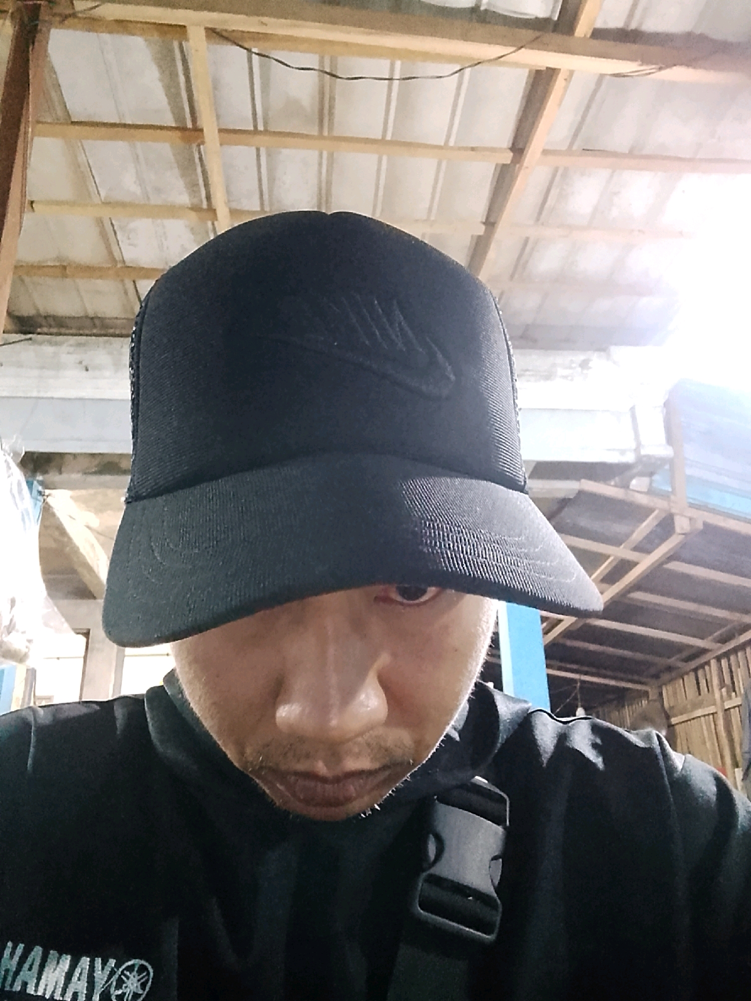 A close-up, selfie-style photo shows a person wearing a black baseball cap with a Nike logo. The person's face is partially obscured by the cap, with only one eye visible. The background suggests an indoor, possibly unfinished or rustic structure with exposed wooden beams and corrugated roofing. The overall lighting is bright, with a strong light source to the right, creating a lens flare effect. The person is wearing a black garment that appears to be a jacket or hoodie, with a black buckle visible on the right shoulder area. Part of a logo, possibly "YAMAHA," is visible on the lower left of the garment. The scene is consistent with a casual indoor setting in Pelaihari, Indonesia, possibly a workshop or storage area.