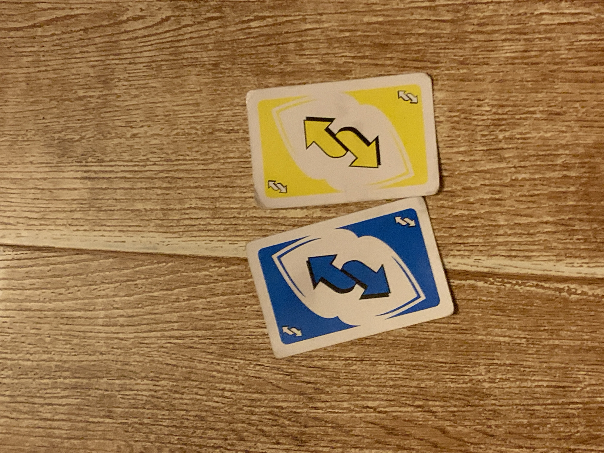 Two Uno reverse cards lie on a wooden surface. One card is blue and the other is yellow. Both cards display the standard Uno reverse symbol: two arrows facing opposite directions in a circular formation. The wooden surface has a visible grain pattern, with vertical lines indicating the wood's texture. The lighting suggests an indoor setting, with no discernible urban or environmental context provided by the image itself. The composition is a close-up shot of the cards.