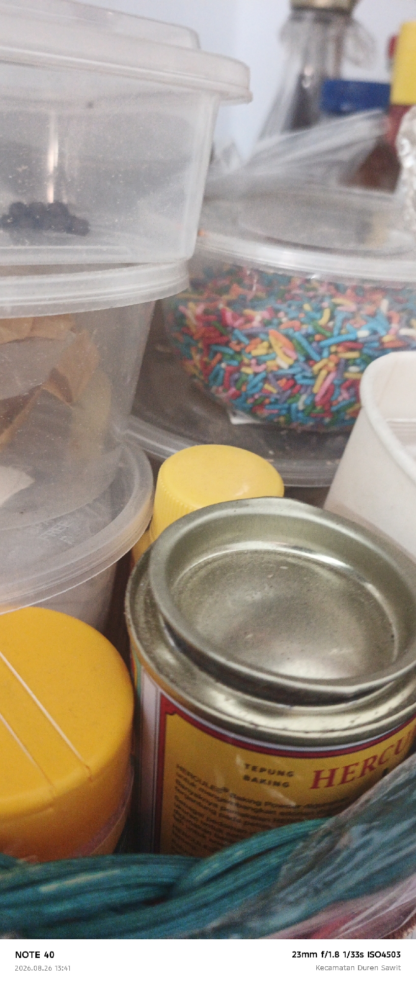 The image shows a close-up, slightly out-of-focus view of various items, likely in a kitchen or storage area. In the foreground, a silver-lidded metal can with a yellow label featuring the word "HERCULUS" and "TEPUNG BAKING" is prominently displayed. To the left of the can, a bright yellow plastic cap or lid is visible. Behind these, a clear plastic container holds colorful sprinkles. Further back, other plastic containers and bottles are visible, some appearing to contain dry goods or ingredients. The background is blurred, with hints of a light-colored wall and other objects. The overall impression is one of everyday items, possibly related to baking or food preparation. The image has a timestamp at the bottom left: "2026.08.26 13:41" and camera details at the bottom right: "NOTE 40 23mm f/1.8 1/33s ISO4503 Kecamatan Duren Sawit". Based on "Kecamatan Duren Sawit", the location is identified as Duren Sawit District, East Jakarta, Indonesia.
