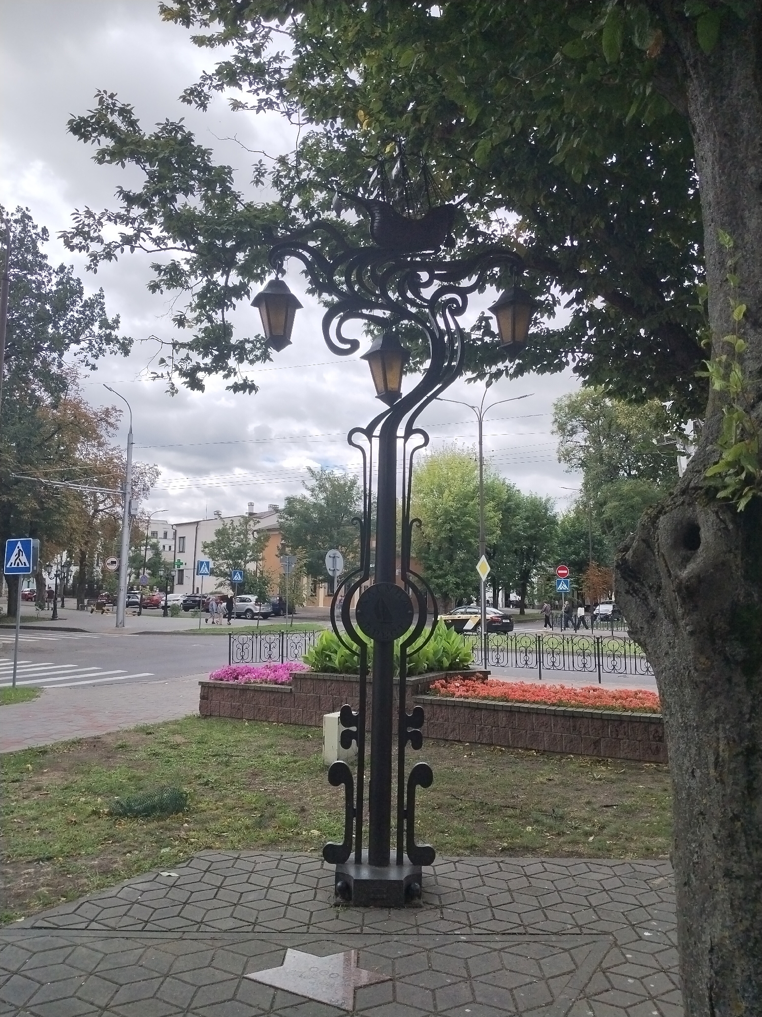 The image depicts a street scene in Brest, Belarus, likely near a pedestrian crossing and intersection. In the foreground, a decorative lamppost with four lanterns is prominently featured, constructed from dark metal with ornate scrollwork. The lamppost stands on a patterned paved area. Behind the lamppost, a low brick planter box contains pink and orange flowers. Beyond the planter, a street with vehicles and pedestrians is visible. Several traffic signs are present, including pedestrian crossing signs and a red circular traffic sign. Buildings line the street in the background, under a cloudy sky. A large tree trunk is visible on the right side of the image, partially obscuring the background. The overall scene suggests a public urban space, possibly a park or square adjacent to a road.