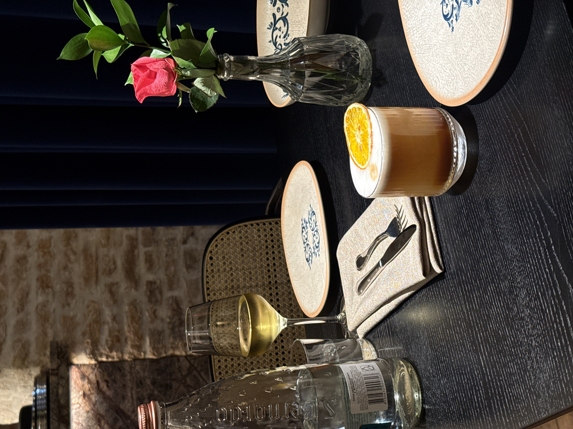 This image presents a well-appointed table setting in what appears to be an upscale indoor restaurant or bar, likely captured during the evening. The dark, textured tabletop is laden with drinks and dinnerware, suggesting a moment before or during a meal.
In the upper left, a single, vibrant pink rose with fresh green leaves is elegantly displayed in a clear, ornate glass vase, set against a backdrop of deep blue drapery. Adjacent to this, a tall, ribbed glass holds a golden-brown cocktail, artfully topped with a layer of white foam and garnished with a dried orange slice, hinting at a sophisticated beverage choice. A distinctive round ceramic plate with a crackled texture and intricate blue patterns is positioned behind the cocktail.
Further along the table, a wine glass filled with a pale yellow liquid, presumably white wine, stands near another identical patterned plate. A neatly folded beige napkin holds a polished silver fork and knife, ready for use. In the foreground, a clear glass bottle of S.Bernardo mineral water is visible, with its label displaying "S.Bernardo" inverted and a barcode: 8009720000431. Additional small text is also present on the label.
The background elements include the woven back of a chair, partially visible behind the wine glass, and a rustic stone or brick wall in the lower left, illuminated by soft, ambient lighting. This combination of elements creates a calm, inviting, and refined atmosphere. No people are visible in the scene.
