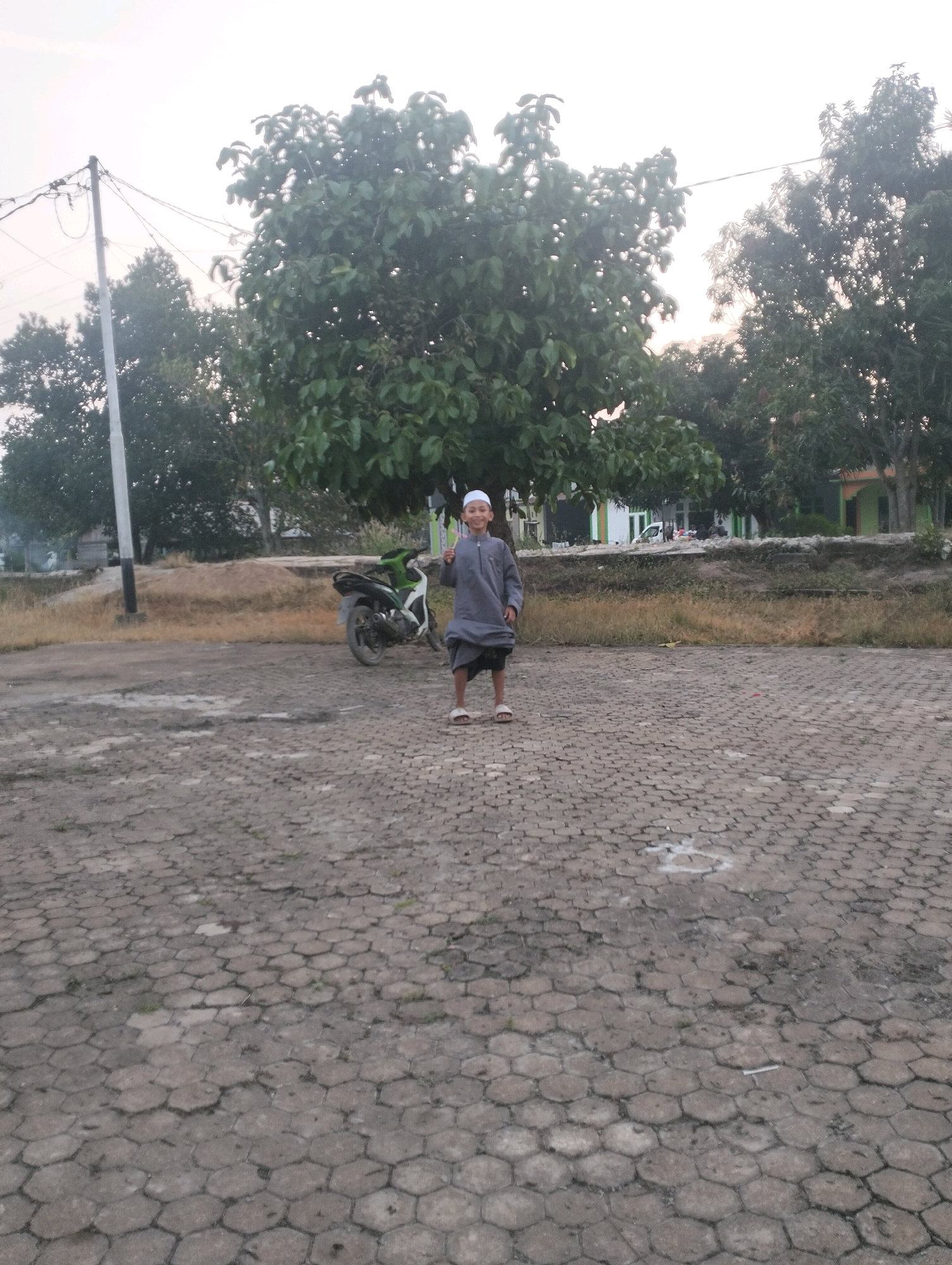 A young boy wearing a white cap and grey traditional attire stands in the middle of a hexagonal paver stone courtyard. He is looking at the camera with a slight smile. Behind him, a motorcycle is parked next to a large tree. In the background, there are more trees and buildings, suggesting an outdoor setting. The sky is bright, indicating it is daytime. The image appears to be taken in a residential area or a public space in Indonesia.