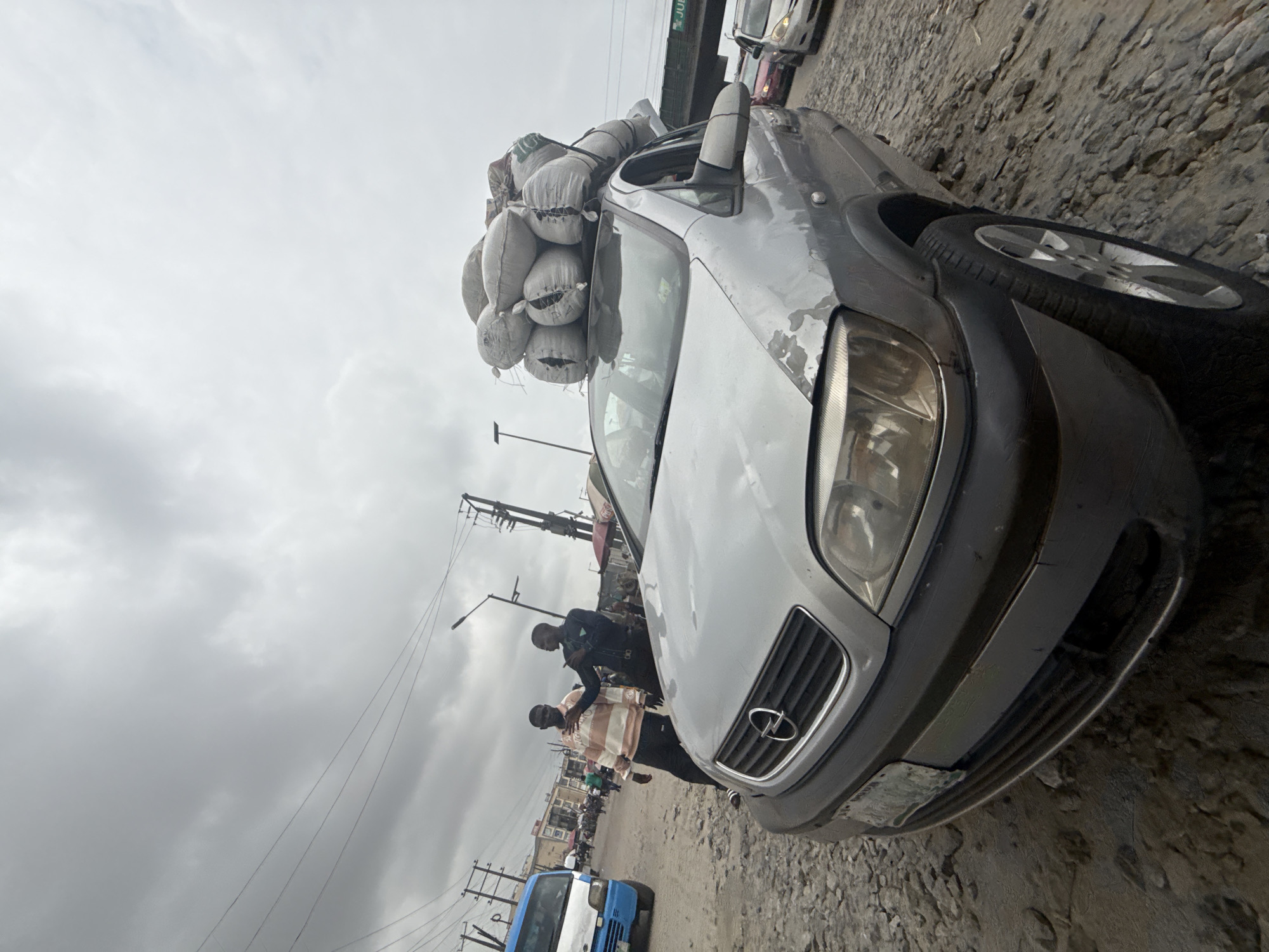 A low-angle, wide shot captures a silver Opel Zafira car parked on a rocky, unpaved road, heavily laden with numerous large, light-colored sacks piled high on its roof. The car's front grille and headlights are visible, along with a portion of its license plate. In the background, a blue and white commercial van is parked to the left. Two men stand near the front of the silver car; one, wearing a striped long-sleeved shirt, gestures with his hands, while the other, in a dark shirt, appears to be speaking. Other people and various vehicles are visible in the blurred background, suggesting a busy street scene. Overhead, a network of power lines stretches across a cloudy, overcast sky. The scene appears to be in Lekki, Nigeria, a coastal area known for its commercial activity and infrastructure development.