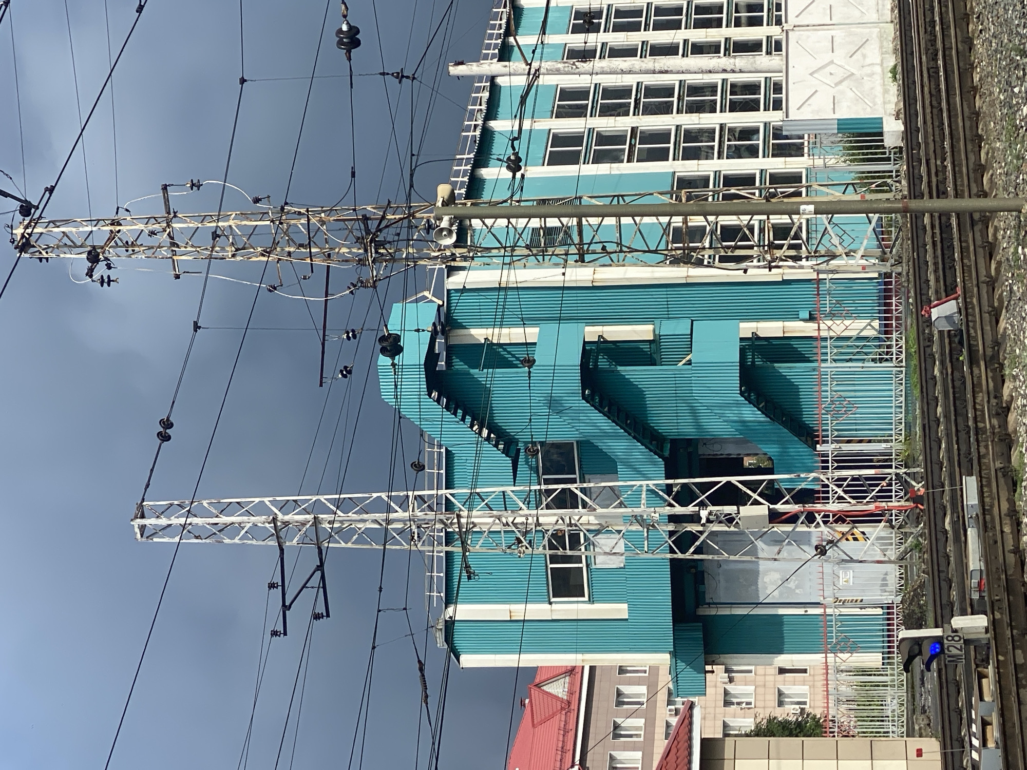 The image depicts an industrial area with train tracks in the foreground. In the midground, a modern teal-colored building with white trim and windows stands prominently, featuring an external staircase. To the left, a light-colored building with a red roof is visible, and further left, another building with multiple windows. Overhead, a complex network of electrical wires and two tall, lattice-style metal poles support them. The sky is overcast with dark clouds. A railway signal displaying a blue light is visible on the tracks in the lower left corner. This scene is located at the Novokuznetsk-Severny railway station in Novokuznetsk, Russia.