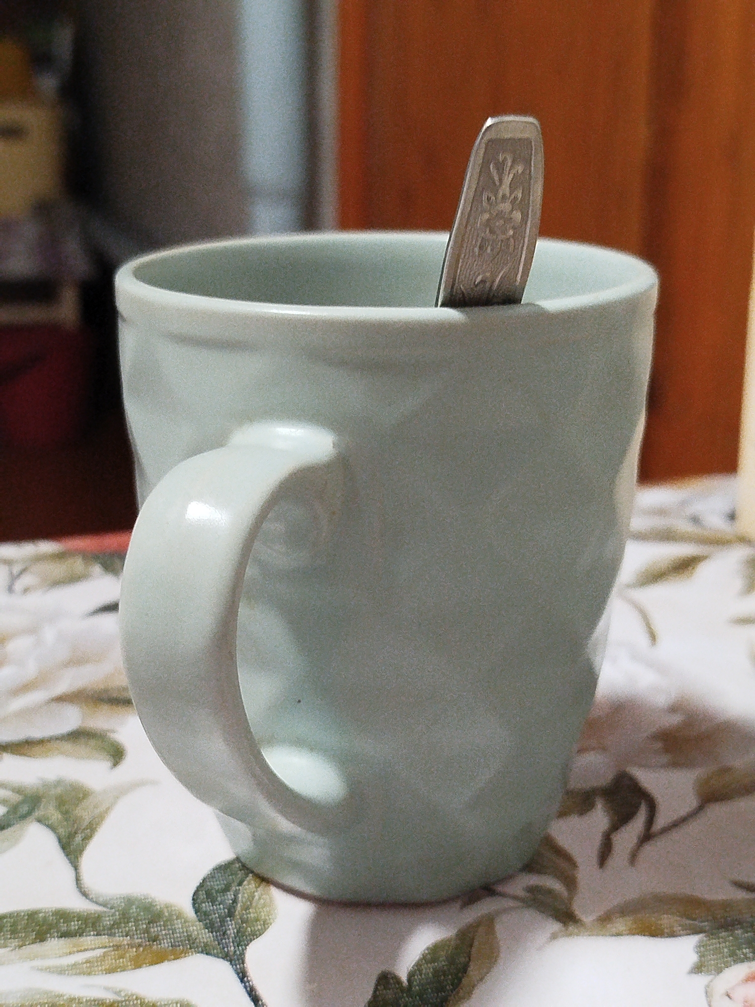 A pale green mug with a spoon inside sits on a floral patterned tablecloth. The mug has a C-shaped handle. The spoon has an ornate design on its handle. The background is blurred, showing a wooden surface and a grey cylindrical object. The scene is indoors and appears to be a domestic setting. The location of Шой-Шудумарь, Russia, is not depicted or implied in the image.
