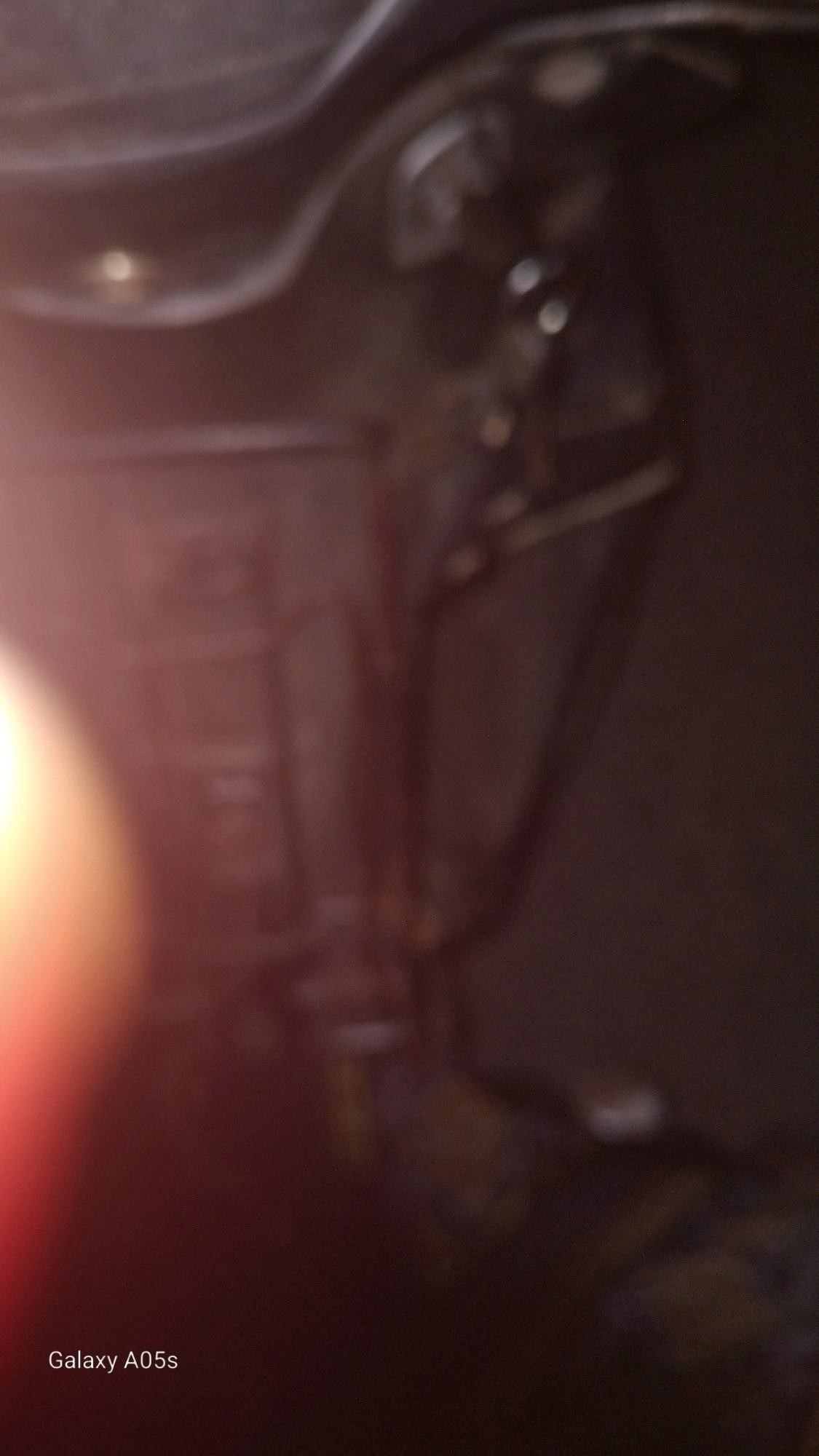 The image is a close-up, blurry shot of what appears to be the underside of a vehicle. There is a strong lens flare on the left side of the image, casting a reddish-orange glow. The background is dark and indistinct, with some lighter areas that could be metal or plastic components of the vehicle. Some circular lights are visible in the upper right portion of the image. The bottom left corner of the image displays the text "Galaxy A05s," indicating the device used to take the photograph. No people or specific environmental details of Sumedang, Indonesia are discernible.