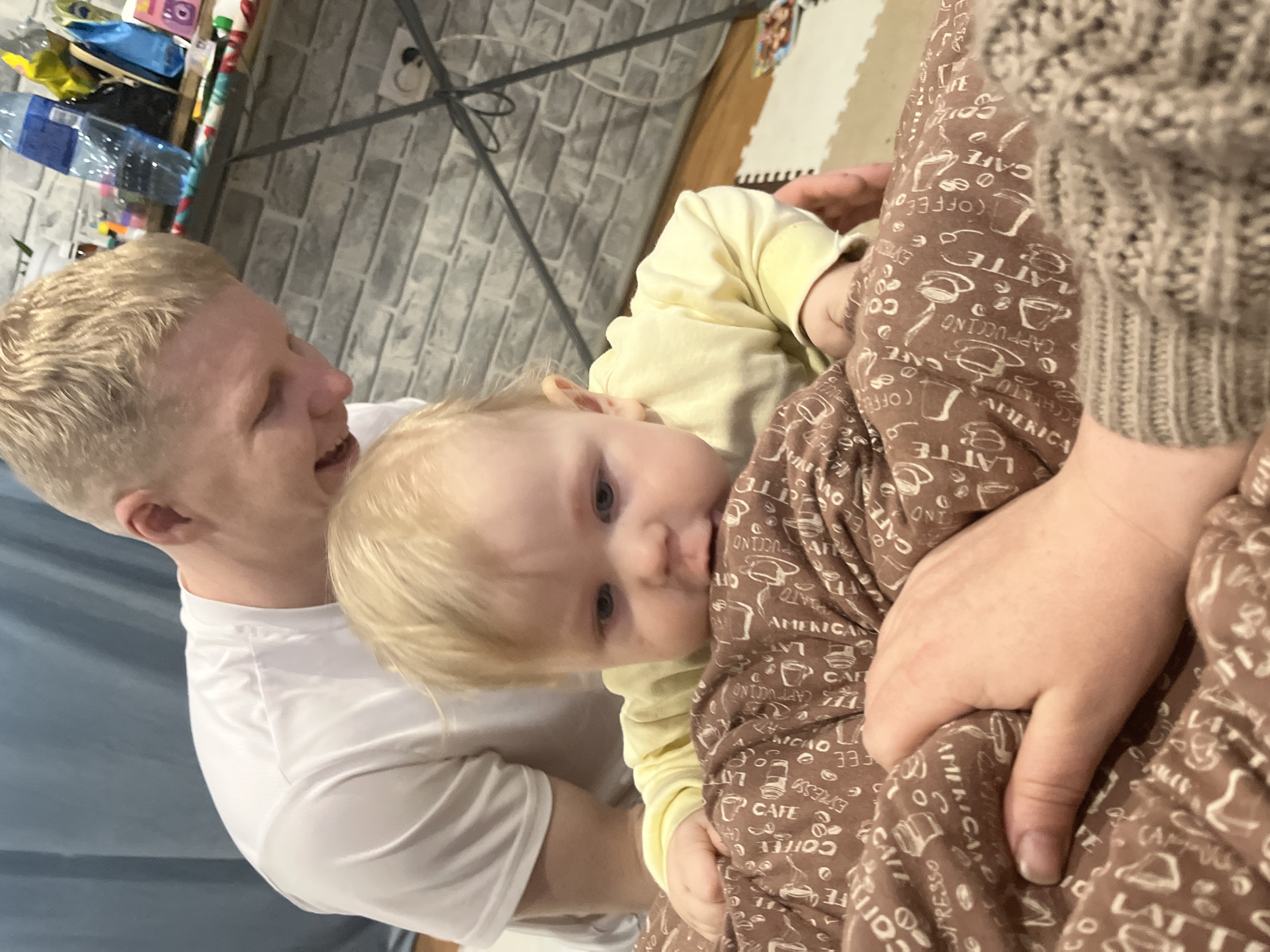 An image captures a tender family moment indoors in Vesyoloye, Russia. A fair-haired man, likely a father, is seen on the left, wearing a white t-shirt. He smiles warmly, looking down at a baby. The baby, also fair-haired with blue eyes and wearing a light yellow long-sleeved top, is held by an adult whose arm and torso are visible on the right. This adult wears a brown knitted sweater and a patterned garment, likely pajamas or a blanket, adorned with coffee-related motifs.
The baby appears curious, looking slightly towards the camera. The man's joyful expression conveys affection for the child. In the background, a gray brick-patterned wall is visible, along with a shelf holding various household items, including a large plastic water bottle with a blue cap. A dark metal frame or support structure is also partially visible against the wall. On the far left, a blue curtain or fabric adds a soft touch to the domestic setting. The floor in the background features a light-colored interlocking foam mat.
Notable text visible on the brown patterned garment includes "LATTE," "CAPPUCCINO," "CAFE," "AMERICANO," "COFFEE," and "ESPRESSO," accompanied by illustrations of coffee cups and beans. The overall scene suggests a comfortable, happy atmosphere within a home.