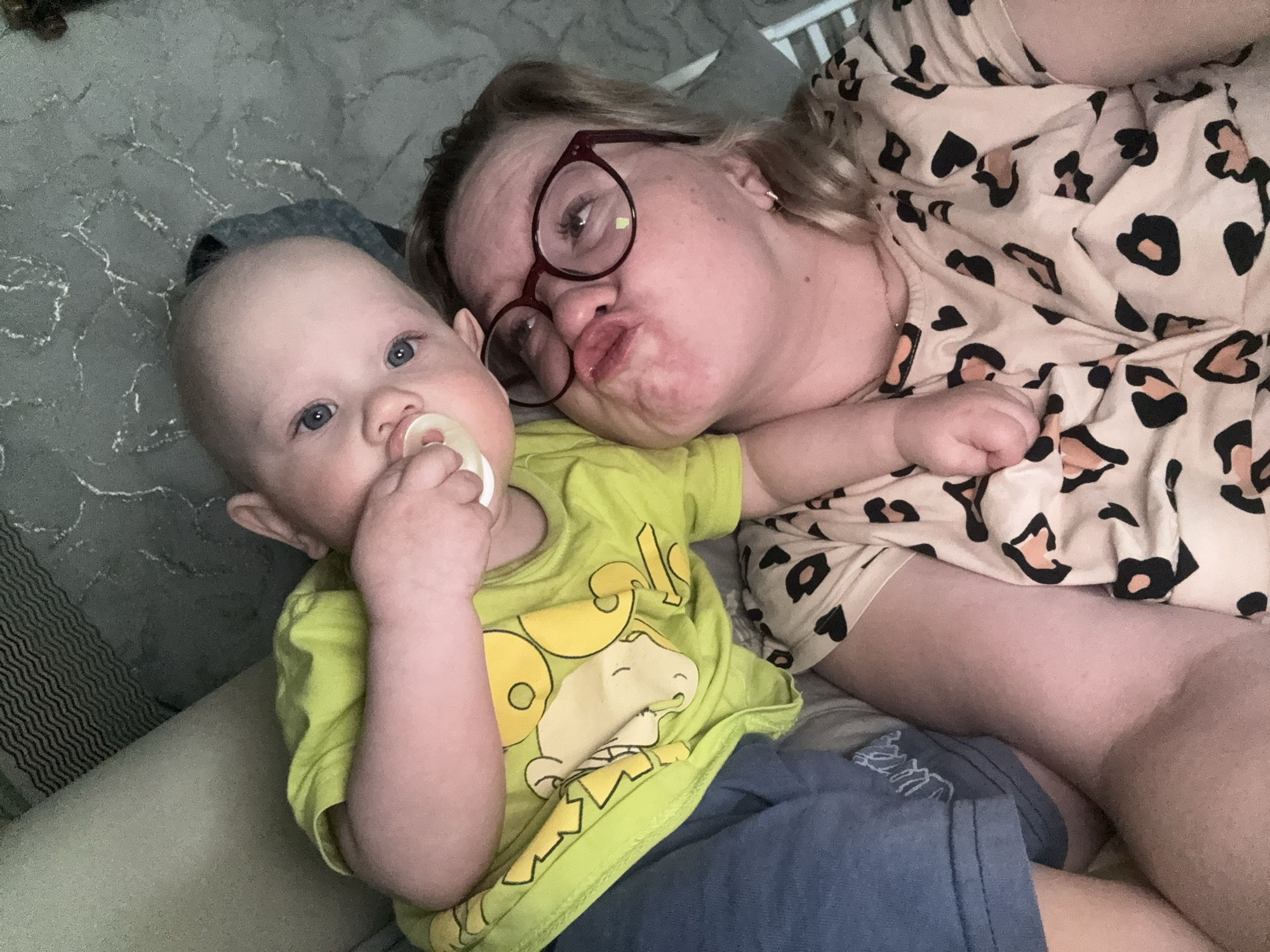 A baby and an adult woman are lying on a textured surface, likely a bed or sofa, in an indoor setting. The baby, with a pacifier in his mouth, is wearing a bright green t-shirt with a cartoon character and blue shorts. The woman is wearing maroon glasses, a patterned beige and black shirt, and is making a kissing face. The environment appears to be casual and intimate. There is no indication of the city of Chaikovsky, Russia.