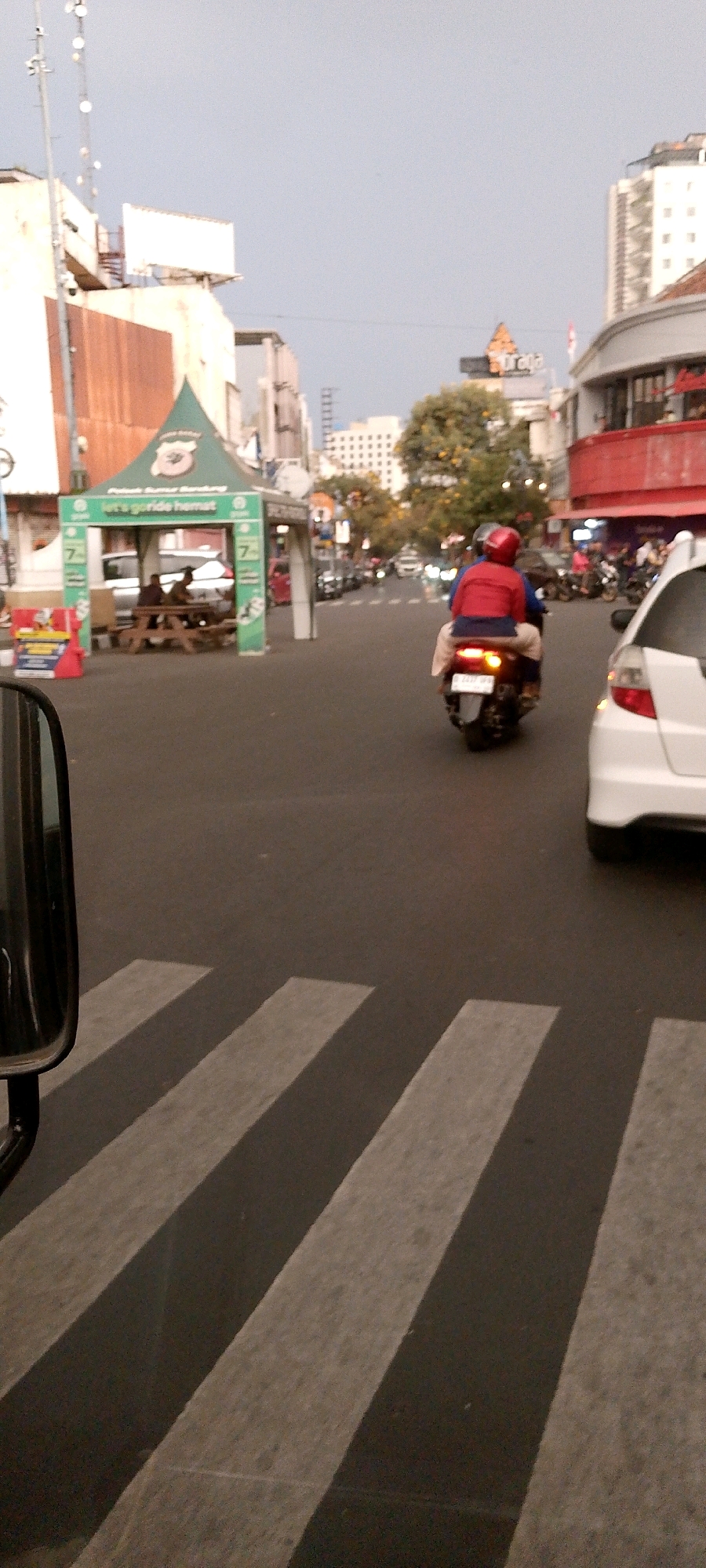 The image depicts a street scene in Bandung City, Indonesia, likely during daylight hours with overcast skies. A pedestrian crossing is prominent in the foreground, with its white stripes partially obscured by the shadow of what appears to be a side mirror from a vehicle. In the middle ground, a motorcycle with two occupants, one wearing a red helmet and jacket, is traveling away from the camera. To the left, a green tent structure with promotional material is set up, featuring picnic tables and people sitting underneath. Buildings of various heights line the street, some with distinctive architectural features. In the background, more traffic and structures can be seen, suggesting a busy urban environment. The overall impression is a snapshot of everyday life in a commercial or pedestrian-heavy area of Bandung.
