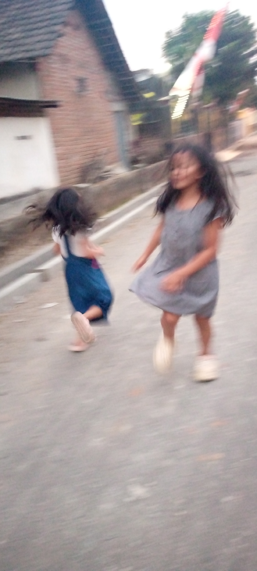 The image shows two young girls running on an asphalt road in what appears to be a residential area in Sumberwono, Indonesia. The girl on the right is wearing a grey dress and the girl on the left is wearing a blue overall dress. Both girls have dark hair and are in motion, suggesting they are playing or running together. The background is blurred, but hints of a building with a brick facade and a tiled roof are visible on the left. A tree with a flag that appears to be the Indonesian flag (red and white) is visible in the upper right corner. The lighting suggests it might be daytime, possibly late afternoon due to the soft light. The overall scene depicts a casual, everyday moment of childhood play in an outdoor environment. There is no visible text in the image.