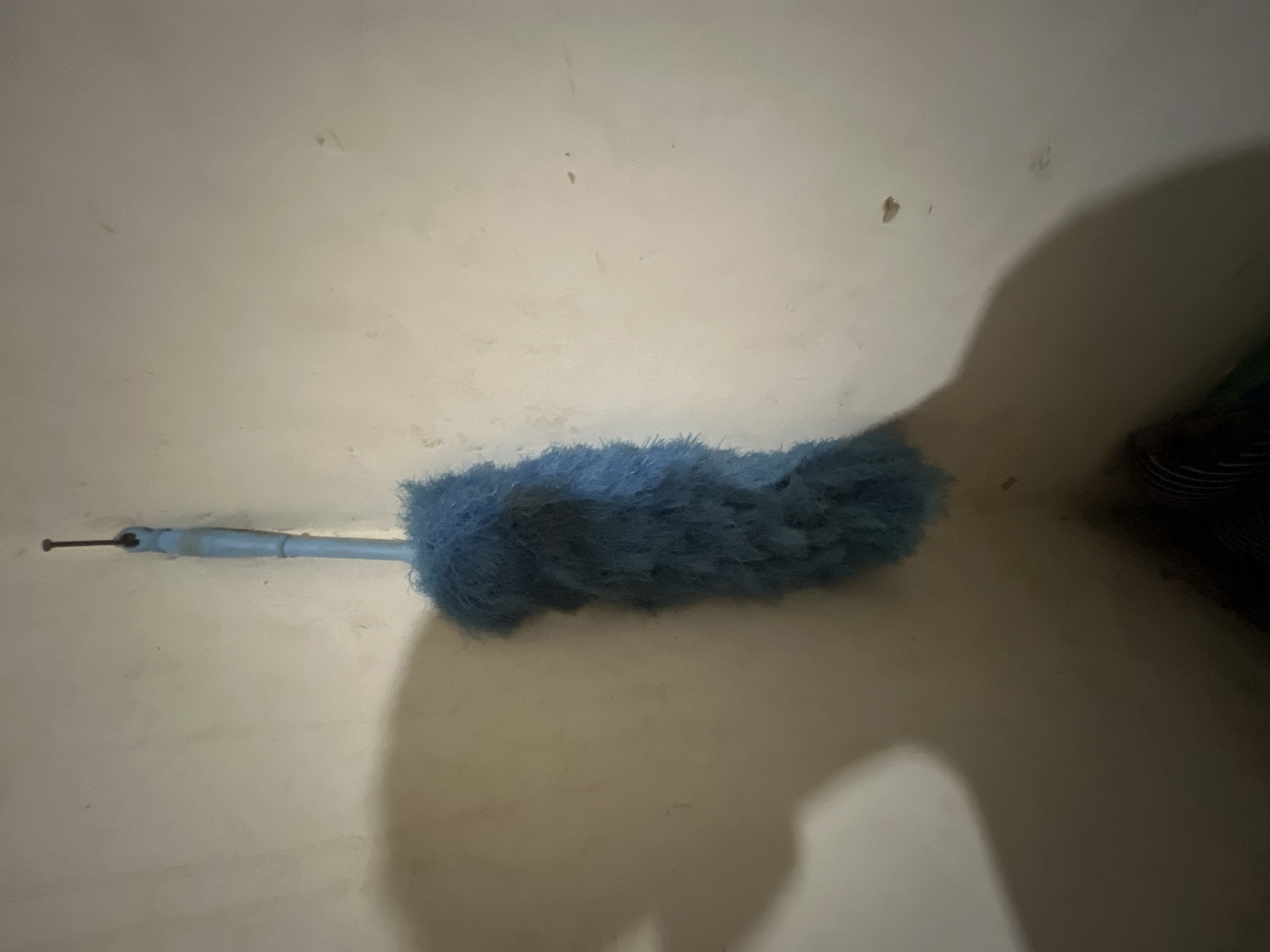 A blue feather duster hangs from a nail in the corner of a room. The walls are a light beige color with some faint marks. A shadow of a person, cast by a light source not visible in the frame, falls across the left wall. The duster's handle is light blue plastic, and the fluffy part is a bright blue. The scene appears to be an indoor setting, possibly a residential or utility space, in Kota Kuala Simpang, Indonesia, as indicated by the context provided.