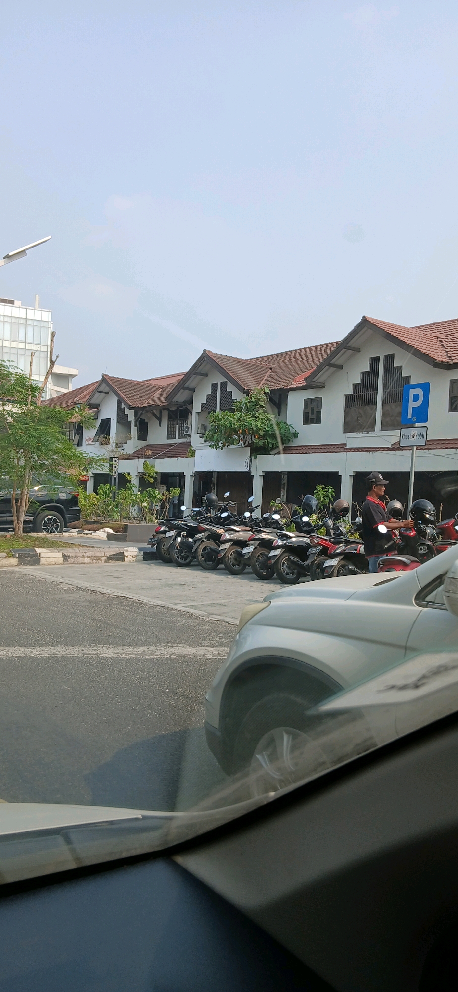 The image depicts a street scene in Samarinda, Indonesia, on a bright, clear day. In the foreground, the hood and windshield of a light-colored car are visible, reflecting the sky and the environment. Beyond the car, a row of motorcycles is parked neatly along a curb. A man wearing a black cap, a red and black shirt, and blue jeans stands next to the motorcycles, appearing to interact with one of them. Behind the motorcycles, a line of white buildings with brown tile roofs stretches across the frame. These buildings have multiple windows and balconies, and some are adorned with green foliage. A blue parking sign with a white "P" is prominently displayed on one of the buildings. To the left of the buildings, a tall modern building with glass windows can be seen, along with a tree with sparse green leaves and a black SUV parked on the side of the road. The street in front of the buildings is paved, with distinct white lines marking parking spaces. The overall environment suggests a commercial or residential area in Samarinda.