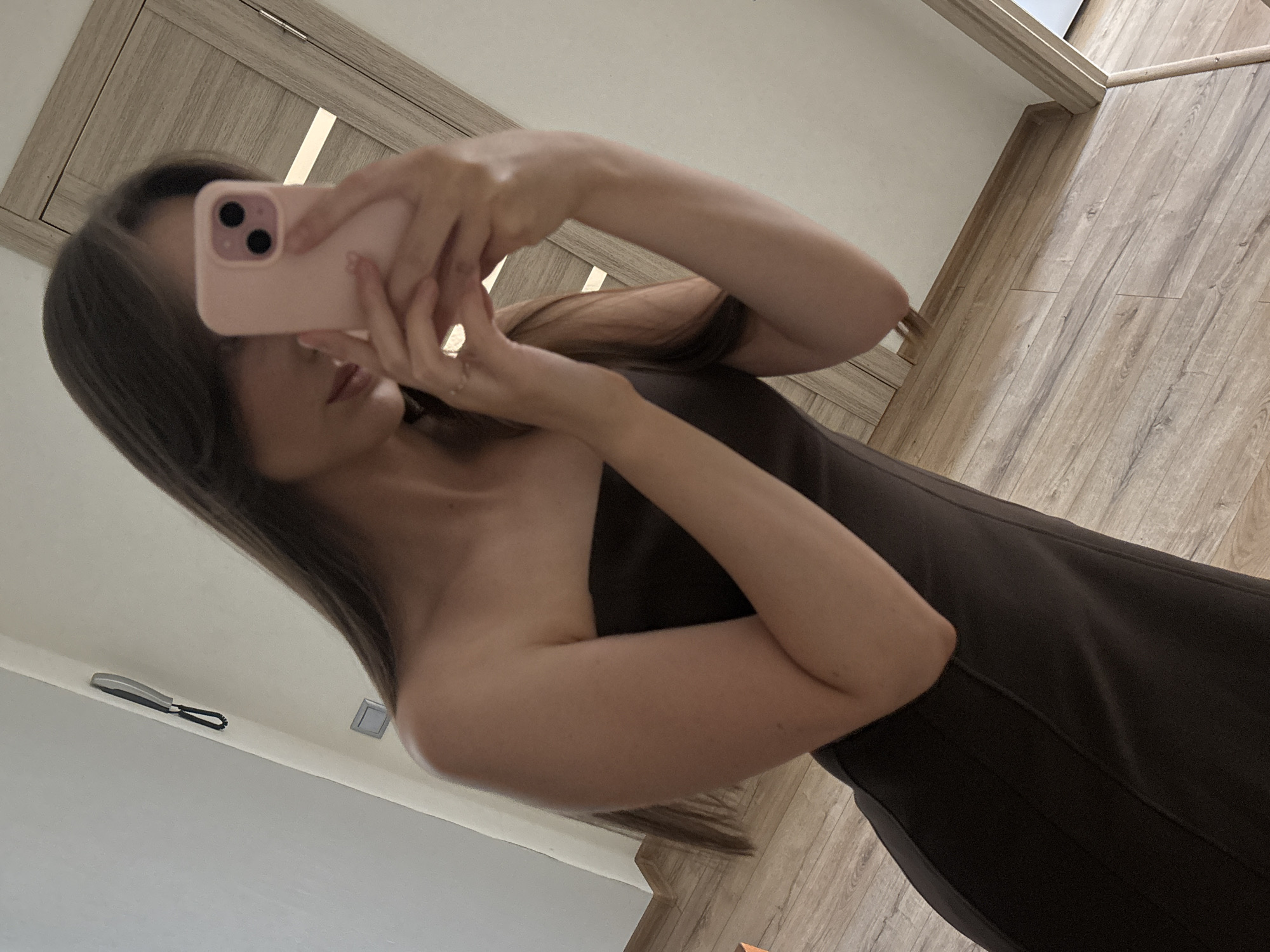 A person is taking a mirror selfie in a room. The person is wearing a strapless brown dress and holding a pink smartphone with two black camera lenses. The person's right arm is bent, and they are wearing a ring on their right ring finger. The background shows a mirror reflecting the person and a wooden door with a white frame. The room has wooden flooring and a white wall. A black and silver intercom-like device is mounted on the wall above a light switch.