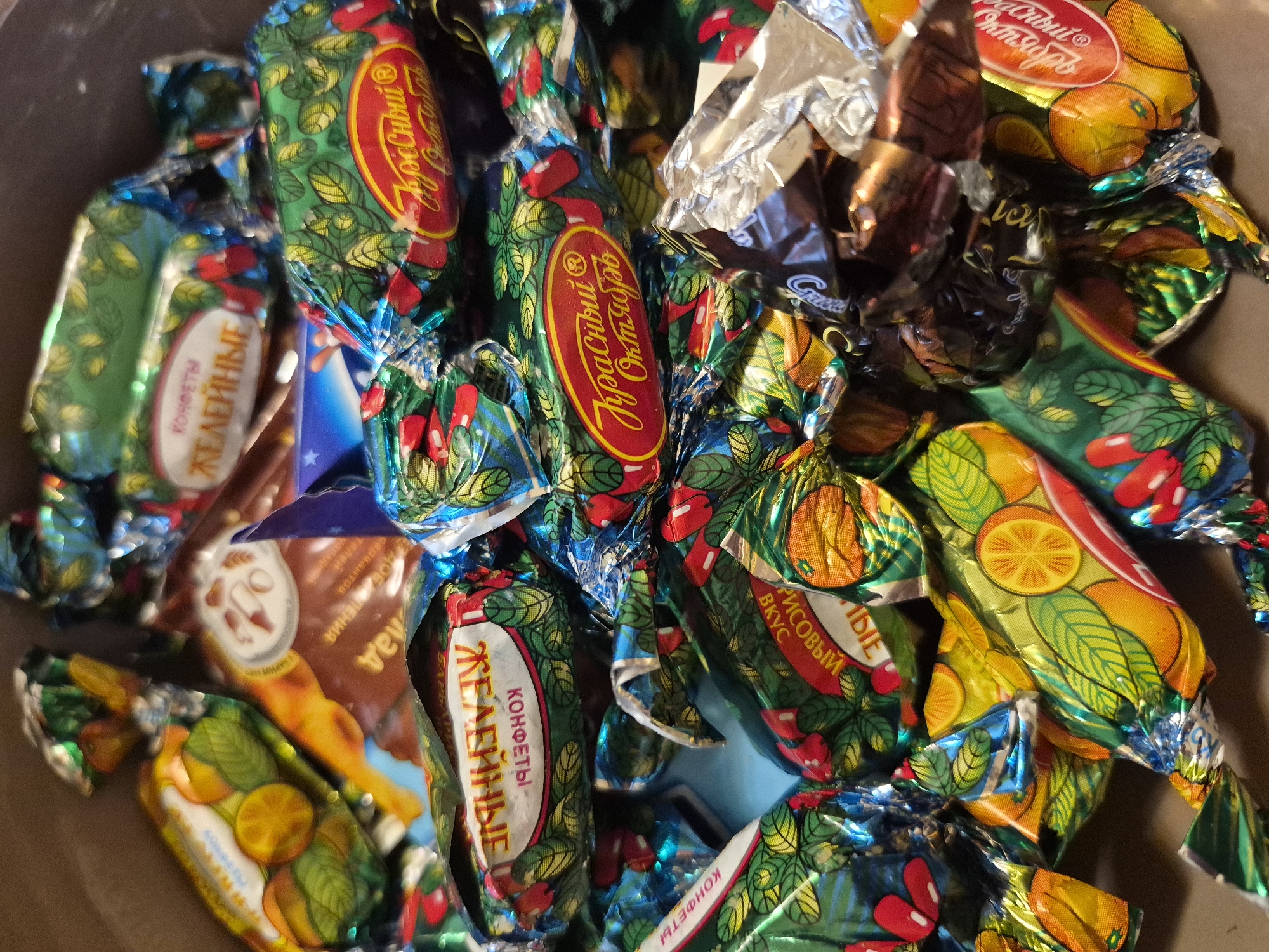 The image shows a close-up, overhead view of a pile of individually wrapped candies, likely in a box or container. The candies have colorful foil wrappers with various designs, including depictions of fruits (oranges and berries) and text in Russian. Some of the wrappers feature patterns of leaves and berries. The lighting is somewhat harsh, creating reflections on the foil. The candies are jumbled together, with some wrappers slightly torn or creased. The background is dark and out of focus. There is no visible indication of a specific location in Yerevan, Armenia, or any people or actions occurring in the scene.