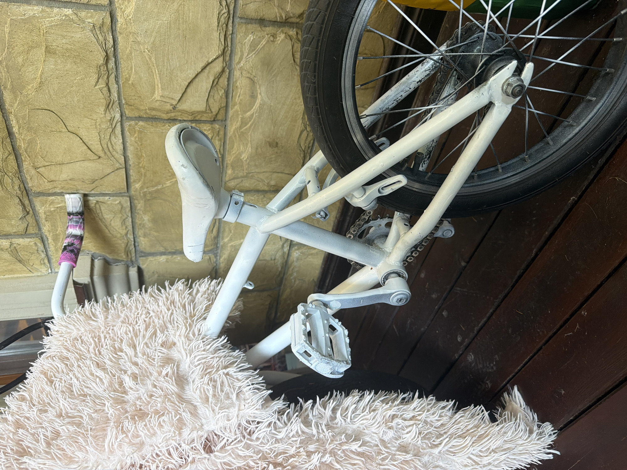 A white BMX bicycle is parked on a dark wooden deck in front of a light brown brick wall. A large, fluffy, cream-colored throw blanket is draped over the handlebars and a portion of the bicycle frame, obscuring part of the front of the bike. The bicycle has white pedals, a white seat, and white frame with some areas of paint chipping. The rear tire is black. The bricks of the wall have a rough, textured surface with visible grout lines. The scene appears to be outdoors. The city of Ленинское, Russia is mentioned as the location, but this information is not visually derivable from the image.