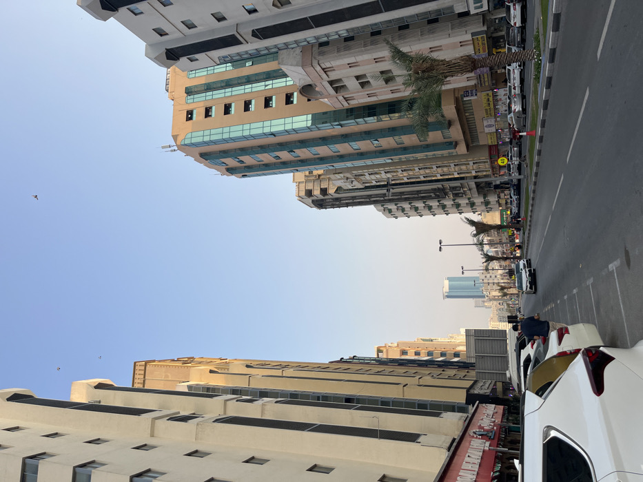 The image displays an urban street scene in the city of Sharjah, United Arab Emirates, looking eastward down a multi-lane road flanked by numerous high-rise buildings. On the left, a row of cream-colored residential or commercial structures rises, with a ground-floor shop featuring a red awning and Arabic script partially visible in the immediate foreground. Several vehicles are parked along the left curb, including a white SUV in the foreground and a yellow sedan adjacent to it. A male individual, wearing a dark blue shirt and dark trousers, stands by the driver's side of the yellow sedan. On the right, similar multi-story buildings line the street, with one prominent structure featuring a light brown facade accented by vertical strips of blue-tinted glass windows. Palm trees are planted along the median strip separating the road from the right-hand sidewalk. Streetlights with multiple fixtures are visible along the road. Traffic, including several white sedans, is observed moving in the distance, and a "40" speed limit sign is visible on the right. The sky is clear and light blue, with several small, dark specks identified as birds. In the far distance, a tall building with a distinctive blue glass facade dominates the cityscape. The lighting suggests late afternoon conditions.