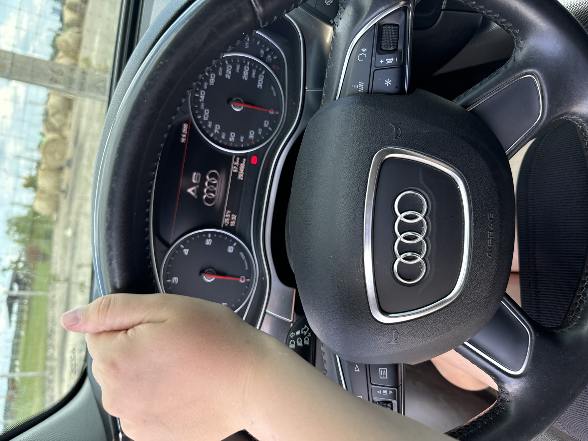 This media file shows a close-up view of a person's hand on the steering wheel of an Audi car. The steering wheel has the Audi logo in the center and controls on the left and right sides, including buttons for navigation and volume. The dashboard is visible, showing a speedometer, odometer, and a digital display with information such as temperature (-25.5°C), time (15:32), date (19.6.2026), fuel efficiency (57.3), and mileage (293400). The car appears to be parked or stopped, as indicated by the static position of the hand and the dashboard readings. Through the windshield, a blurred outdoor scene with what looks like construction materials or hay bales and a clear sky can be seen, suggesting it is daytime with good weather. The overall impression is that of someone inside a car, possibly taking a break or preparing to drive. The visible text includes the car's model "A6" and various dashboard indicators. The location context provided is Kostiantynivka, Ukraine.