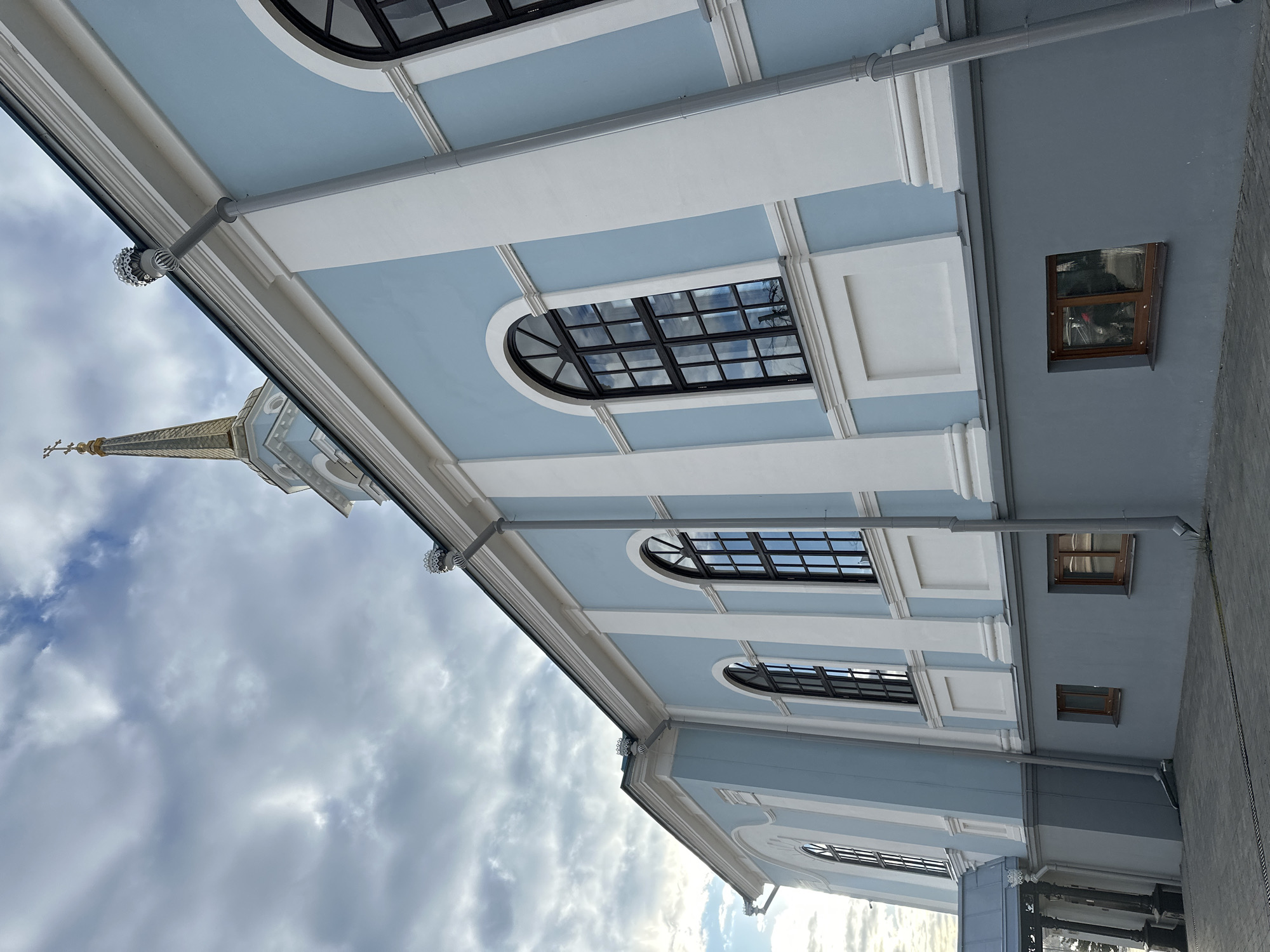 The image depicts a section of architecture in Votkinsk, Russia. The focus is on the exterior of a light-blue building with white architectural details, possibly a church or cathedral judging by the spire partially visible in the upper-left of the frame. The building has arched windows with black frames. The lighting suggests daytime with cloudy skies. There is a gray gutter system running along the edge of the building. The image is shot from a low angle, emphasizing the height of the building. A small window is situated on the right side of the building.