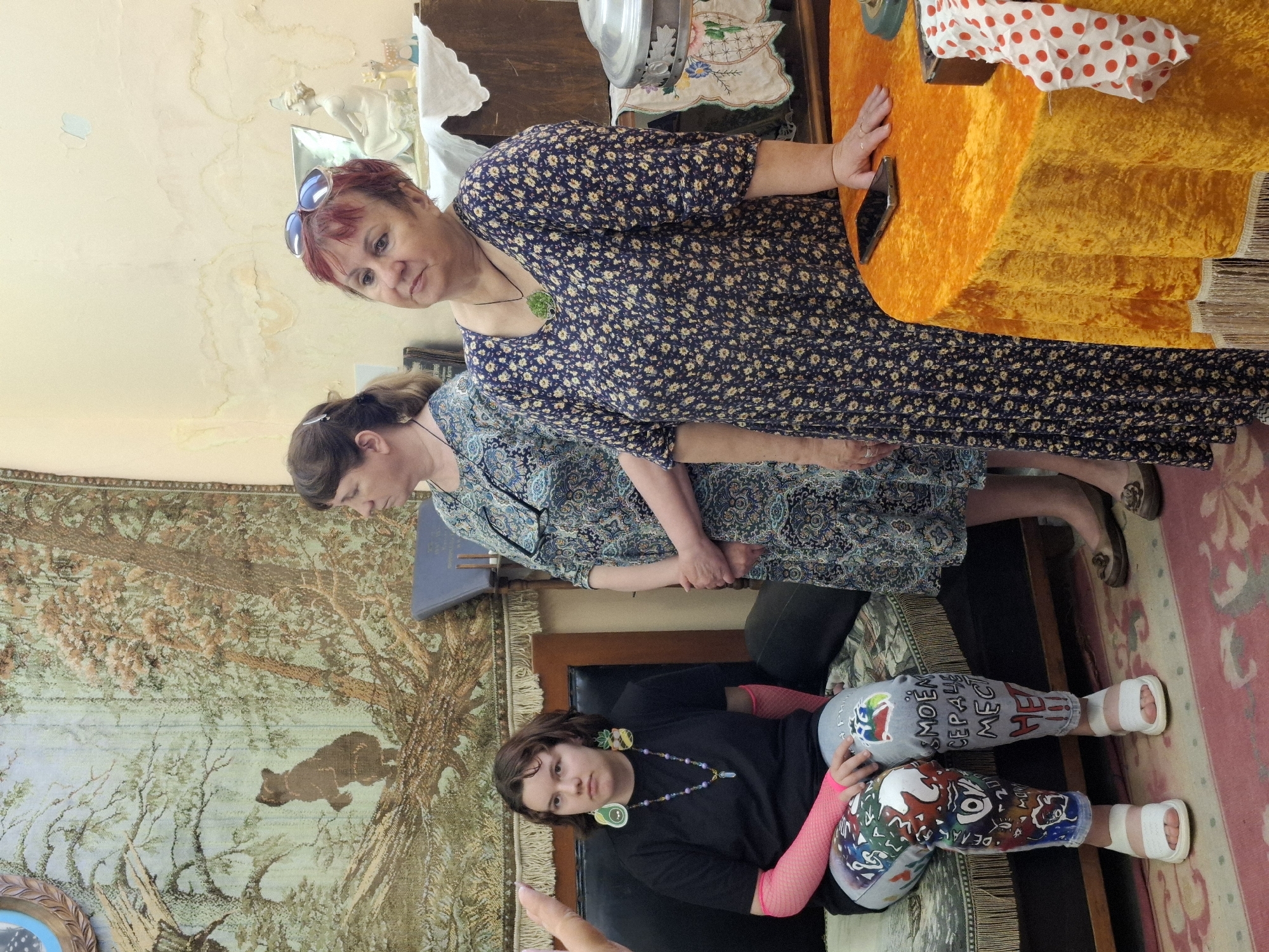 The photograph, taken in what appears to be a residential setting, possibly in Марковский, Russia, shows three women of varying ages. The foreground features a young woman seated on a black leather-like chair. She is wearing a black shirt, patterned leggings, and white sandals with prominent bandaging on her feet. Pink fishnet arm warmers adorn her forearms. She has on a long necklace and fruit-shaped earrings. Behind her stands a middle-aged woman in a blue patterned dress with closed-toe shoes. An older woman, also wearing a blue floral-patterned dress, is positioned at the top of the frame leaning against a golden table. Her short red hair is visible, and she wears sunglasses on top of her head.
The room includes a tapestry depicting a forest scene, and there are various other furniture pieces and decor elements visible. There is an old dresser behind the older woman. The flooring appears to be covered with a patterned rug. There is an orange-yellow table or shelf in the upper right of the image. The lighting appears to be natural, with some shadows and reflections.