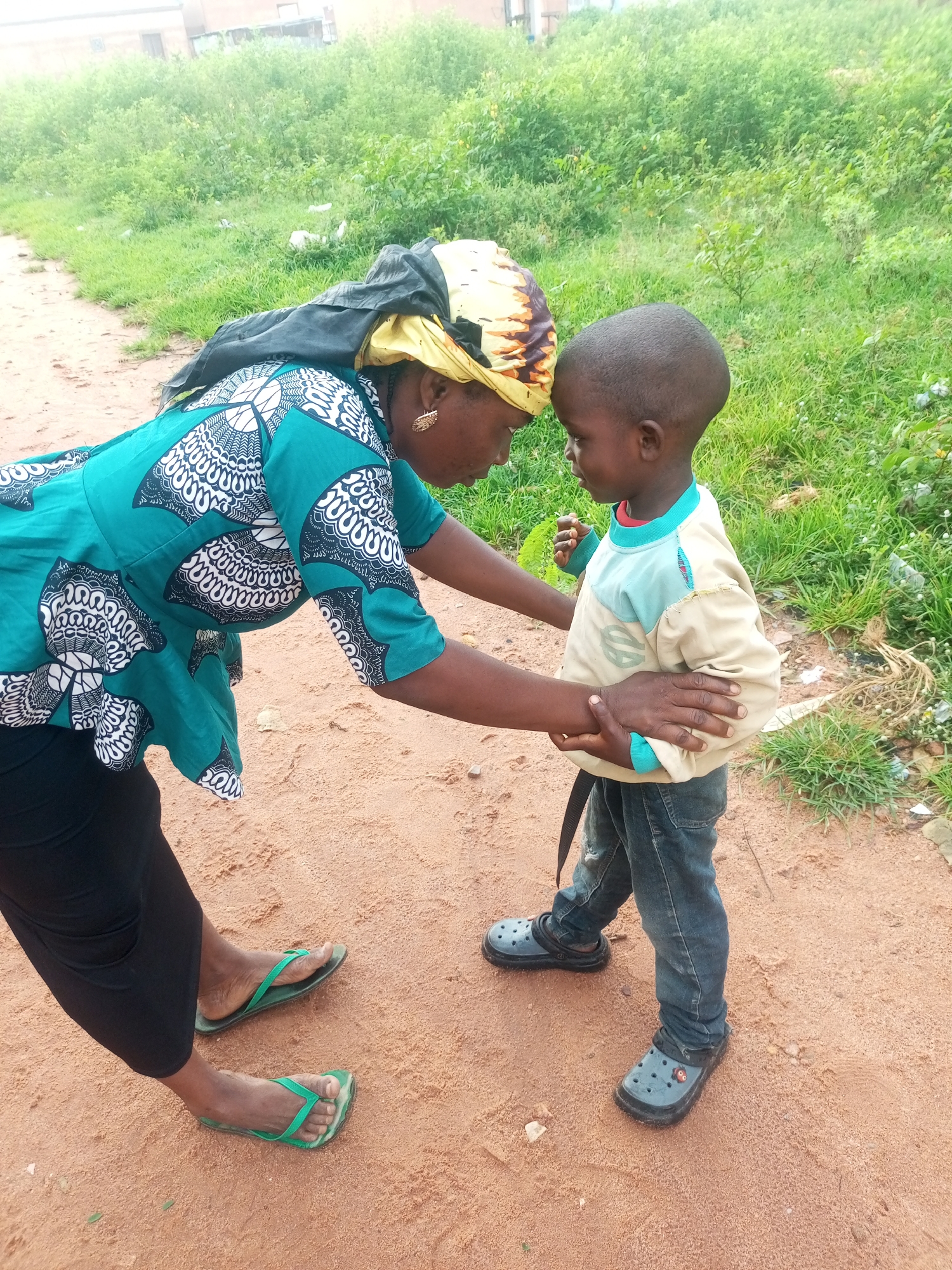 A woman and a young child are standing on a dirt path in Tempure, Nigeria, in front of a patch of tall green grass and vegetation. The woman, wearing a teal and white patterned top, black pants, and green flip-flops, leans down to interact with the child. She has a yellow and patterned headscarf and large gold earrings. The child, wearing a tan and light blue long-sleeved shirt and denim jeans with gray clogs, stands facing the woman. The woman has her left arm around the child's waist and her right hand resting on the child's chest. The child's right hand is touching the woman's hand. In the background, there are some buildings and more vegetation. The ground is made of reddish-brown dirt with a few small pebbles.