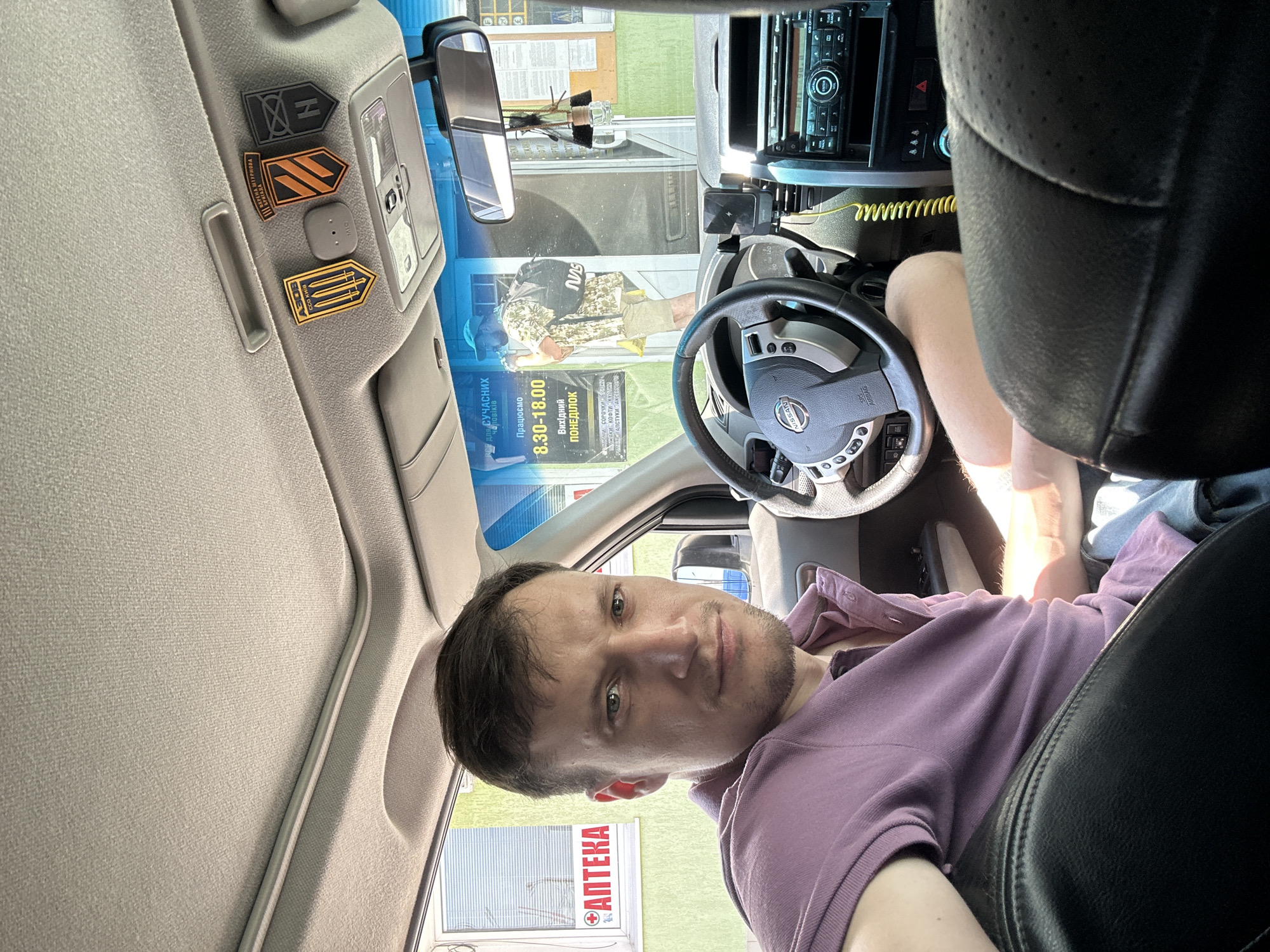 The image shows a man in the driver's seat of a car, looking towards the camera. He is wearing a purple polo shirt. The interior of the car, including the dashboard and steering wheel, is visible. Outside the car, through the windshield, a building with a sign that reads "АПТЕКА" (pharmacy) can be seen. There is also a poster with text in Ukrainian. The overall scene suggests the man is stopped at a location likely in or near the city of Kamianets-Podilskyi, Ukraine, possibly at a drive-through or service point.