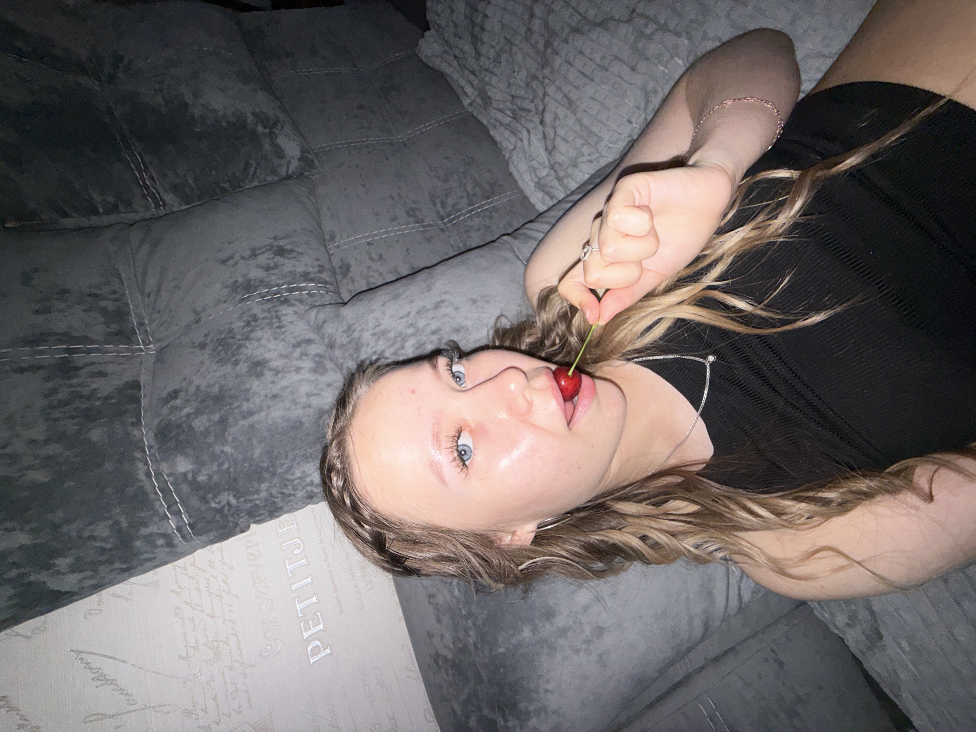 A young person with blonde hair and blue eyes is lying on a gray couch, holding a cherry with a green stem to their mouth. They are wearing a black top and a silver necklace. Their hair is long and wavy, with a braid on the left side of their head. The couch has a textured surface and white stitching details. In the background, another part of the couch is visible, covered in a textured gray fabric. A section of the couch has a light-colored surface with decorative text and patterns, including the word "PETITJE" in large capital letters. The lighting is dim, suggesting an indoor setting, possibly in the evening or a dimly lit room. The person's expression is playful or alluring, with their eyes looking towards the camera and their lips slightly parted around the cherry. There are no explicit clues about the time of day, weather, or specific location within Chaikovsky, Russia, beyond the general indoor setting.