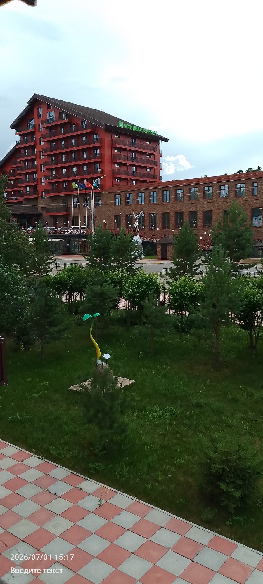 The image depicts a view of the Wyndham Garden hotel in Burabay, Kazakhstan. In the foreground, a red and white checkered tiled pathway is visible. Beyond the pathway is a lush green lawn with trees and a quirky sculpture of a sprouting plant, which is yellow with green leaves. In the mid-ground, there is a multi-story brick building with numerous windows and a wooden balcony structure. In front of this building, flags are flying, and a large metal sculpture of a deer stands on a pedestal. The background features a tall, red-toned hotel building with many balconies and windows. The sky is overcast.