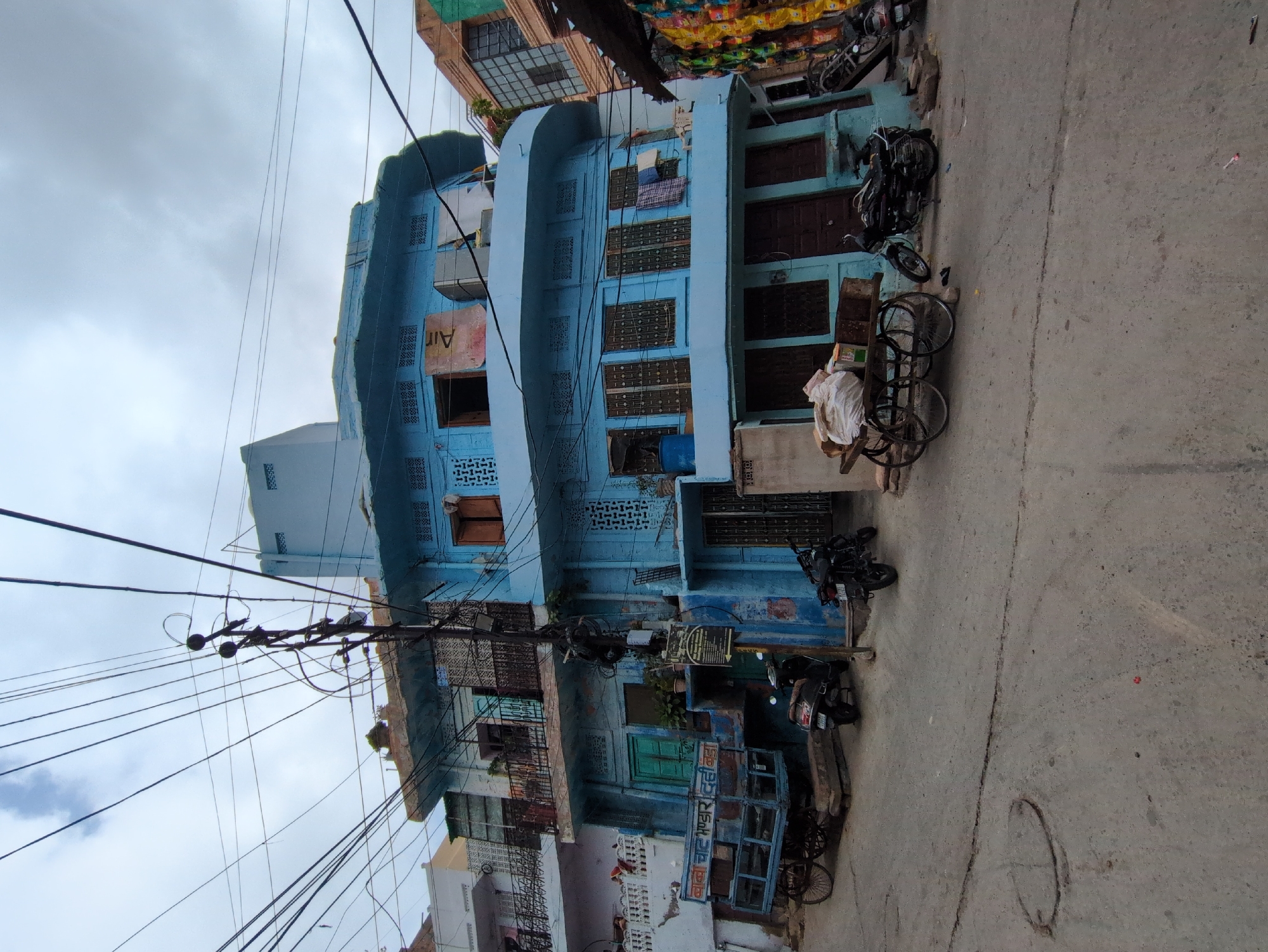 The image shows a street scene in Jodhpur, India, with a distinctive light blue building dominating the frame. The building has multiple stories, balconies with intricate metal railings, and a curved architectural element on the upper floor. Numerous electrical wires crisscross overhead, strung between a utility pole and various structures.
In front of the building, a blue cart with wooden sides and spoked wheels is parked. It appears to be a type of handcart or pushcart, possibly used for transporting goods. Several motorcycles are parked on the paved street, including two dark-colored scooters and a motorcycle with a rider in dark clothing.
The street is paved with concrete or asphalt and shows some signs of wear, with a visible crack running across it. A round metal manhole cover is also present on the street. The buildings on either side of the blue structure appear to be of similar architecture, with ornate windows and balconies. Some laundry is visible hanging from a balcony of the blue building. Bags of what appear to be groceries or merchandise are stacked on a wooden cart in the foreground. The sky is overcast with grey clouds.