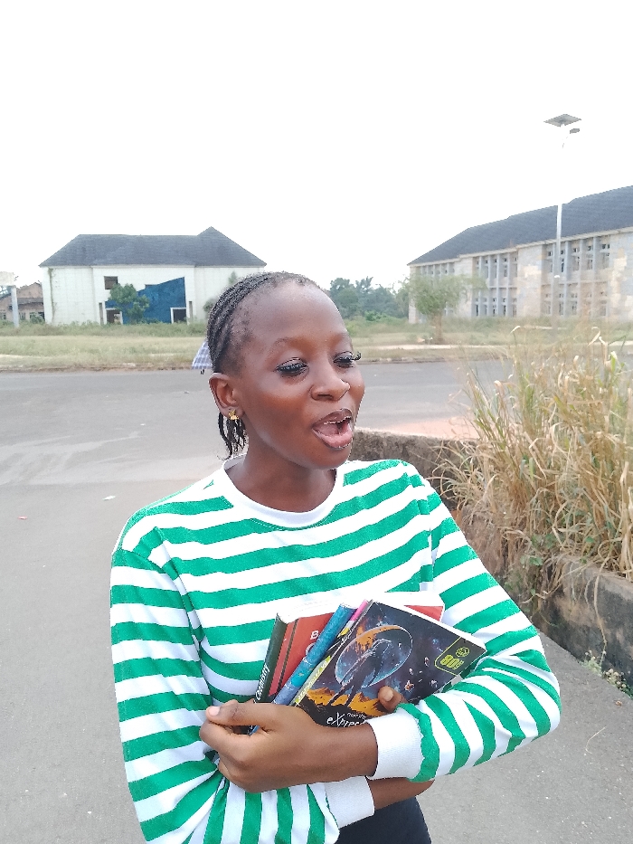 A young Black female stands outdoors in Umueshi, Nigeria, holding a stack of books with her left arm. She wears a long-sleeved shirt with horizontal green and white stripes and has braided hair. Her mouth is open, suggesting she is speaking or reacting, and she looks towards the right of the frame. In the background, a paved road separates her from two distinct buildings. To the left, a single-story white building with a dark roof shows a large blue tarp covering a section. To the right, a multi-story, light-colored building with multiple windows stands. Sparse, dry vegetation is present along the road's edge to the right, where a street light with a solar panel is also visible. The sky is bright and overcast.