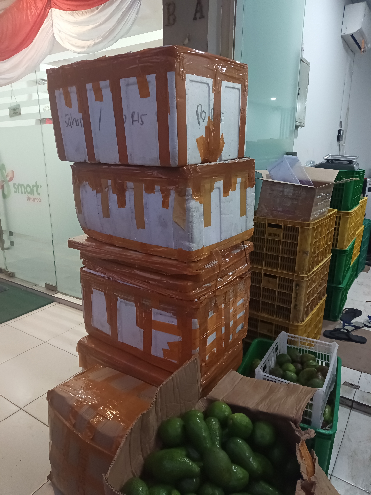 In North Jakarta, Indonesia, an indoor scene depicts a collection of stacked items, likely prepared for shipping or storage. In the foreground, a cardboard box is filled with ripe green avocados, some spilling out onto a white plastic crate also containing avocados. Behind the box, several large styrofoam coolers are stacked vertically, wrapped in brown packaging tape. Some of these coolers have white labels with handwritten text, including "Sahan" and "PONS". Adjacent to the coolers are stacks of yellow and green plastic crates, typical for transporting produce. A digital scale is visible in the background. The environment appears to be a commercial space, with a glass door to the left featuring a "TUTUP" (Closed) sign and the "smart finance" logo. Red and white fabric is draped overhead, suggesting a decorative or celebratory element. The lighting is bright, indicating an indoor setting.