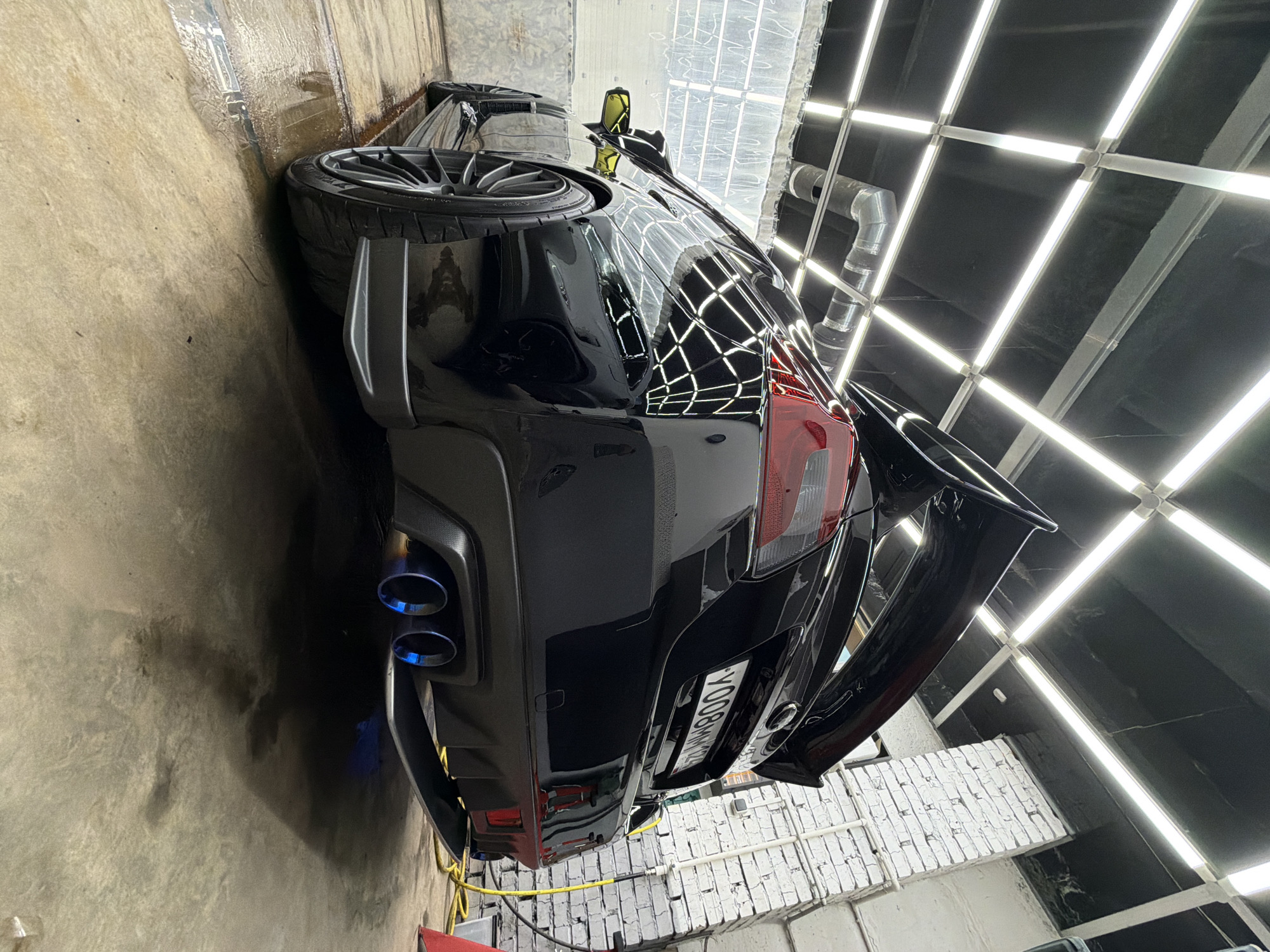 The image displays the rear-right side of a black performance car, likely a sports sedan, positioned vertically in what appears to be an indoor car detailing or wash bay. The car's highly reflective black paintwork mirrors a grid of bright white rectangular ceiling lights, characteristic of a professional detailing environment.
Notable features of the car include dual exhaust pipes with striking blue-tinted tips and a prominent rear spoiler. A Russian license plate, "Y008MM 124," is affixed to the back, with "124" indicating the Krasnoyarsk Krai region. The car's right rear wheel, equipped with a multi-spoke rim, is also visible. The surrounding area features a wet concrete floor and walls constructed from both raw concrete and white-painted brickwork. A yellow hose or cable is coiled on the ground near the vehicle. Overhead, a large, silver, ribbed pipe, likely part of a ventilation system, runs along the ceiling next to the artificial lighting. The scene suggests the car has recently undergone washing or detailing.