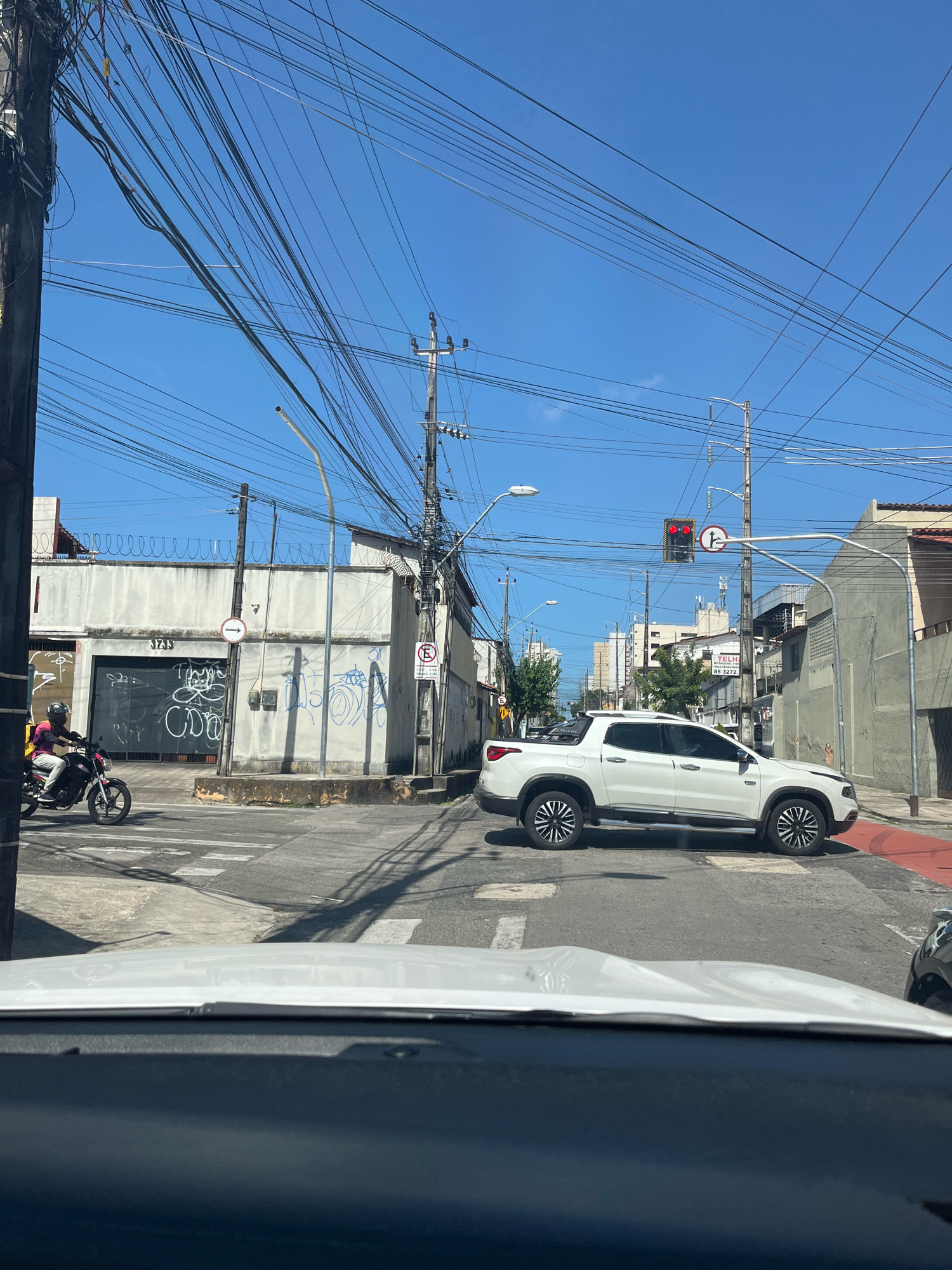 The image shows a street scene in Fortaleza, Brazil, under a clear blue sky with numerous overhead electrical wires. A white pickup truck is stopped at an intersection, its tires positioned on a red-painted curb. To the left, a person on a motorcycle wearing a helmet and a pink shirt is also at the intersection. Behind the motorcycle, a building with a large garage door covered in graffiti is visible, along with a street sign indicating a one-way direction. Further down the street, there are more buildings, some with awnings and visible windows. Traffic signals are present, with one showing a red light. The foreground is dominated by the dashboard and windshield of a vehicle, indicating the photo was taken from inside a car.