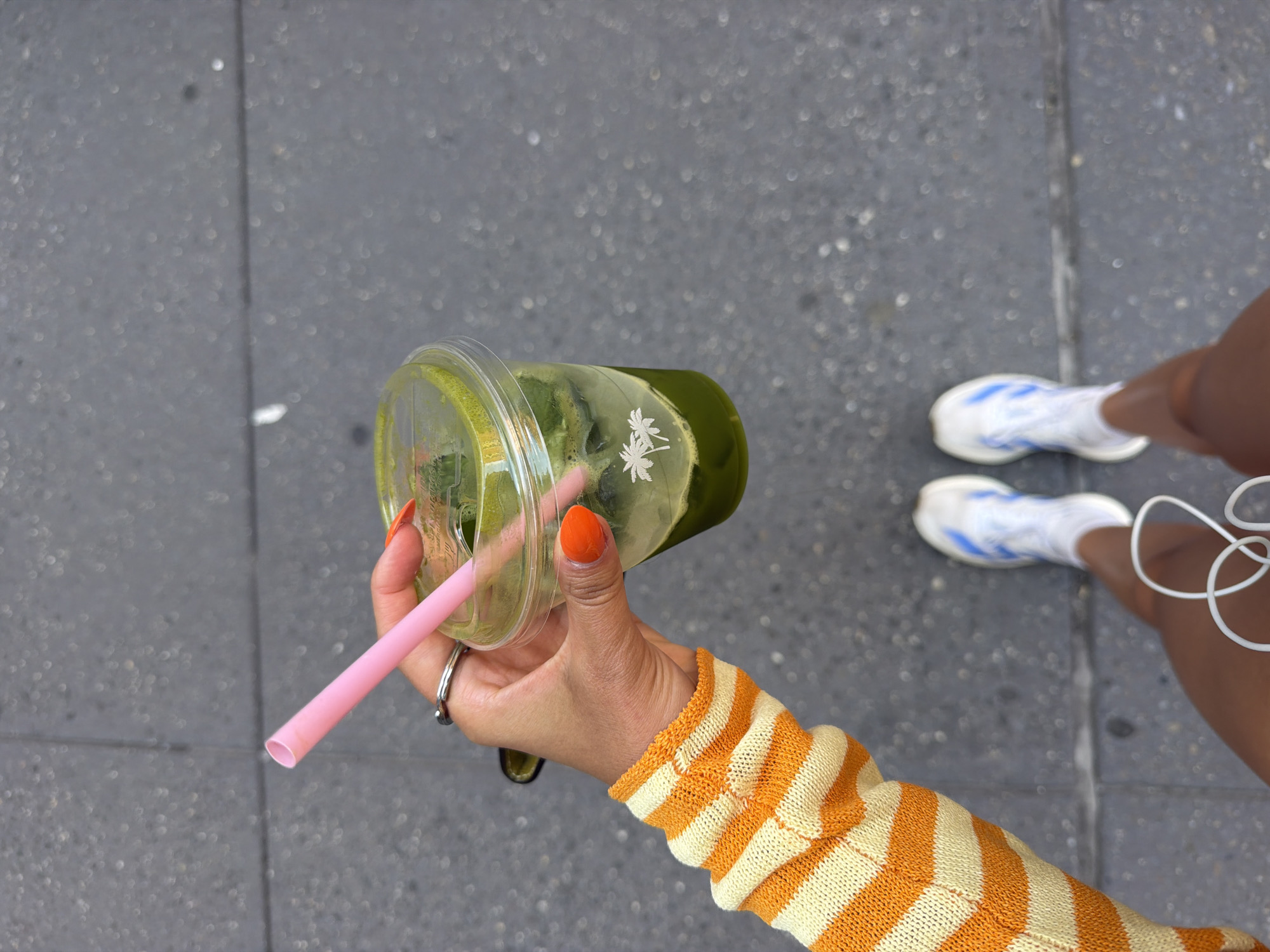 An aerial shot depicts a person holding a transparent plastic cup filled with green liquid. The cup has a lid and a pink straw inserted. The cup features a palm tree logo. The person's hand has orange nail polish and wears a ring. They are wearing a yellow and orange striped sweater. The ground is gray with a mottled texture. The lower part of the frame shows the person's legs, with white sneakers and blue stripes. A white cord, possibly from earbuds, is also visible near the legs. The location appears to be an outdoor urban setting, potentially a sidewalk. It may be in the city of New York, United States of America.