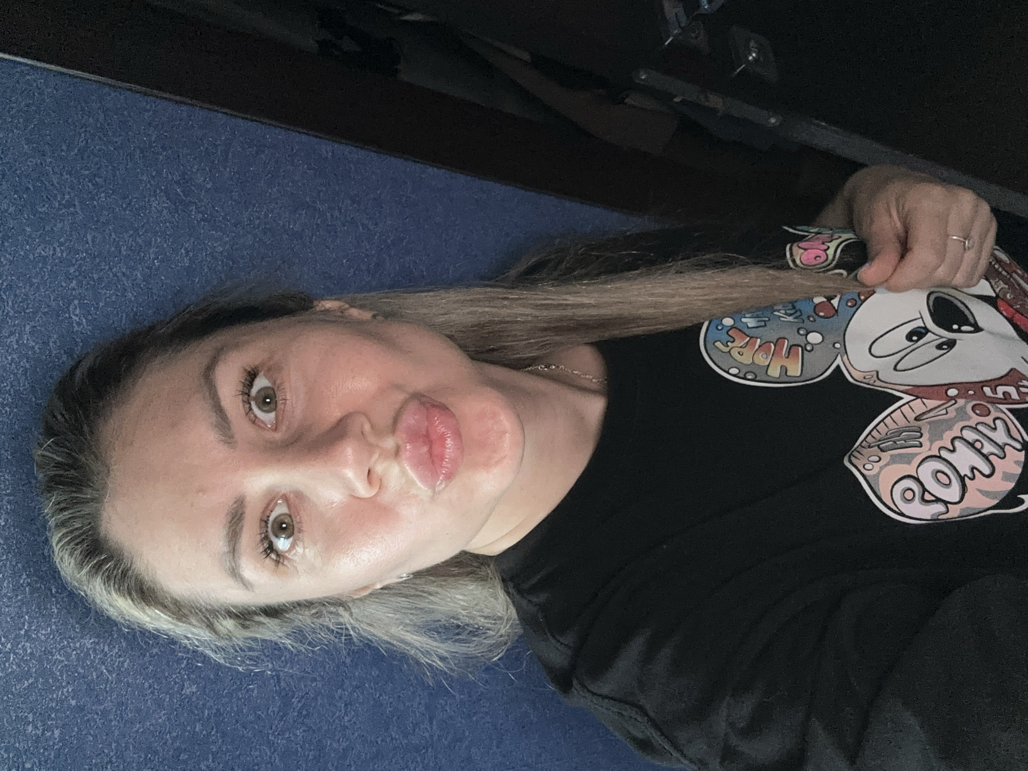 The image shows a selfie of a woman with blonde hair and light brown eyes. She is wearing a black t-shirt with a graphic print of Mickey Mouse on it. Her lips are pursed in a kissing gesture. She is standing in front of a textured blue wall. To the right of the woman, a dark, indistinct object is visible, possibly furniture or a doorway. The lighting is dim, suggesting an indoor setting. The location is identified as Novoaltaysk, Russia.