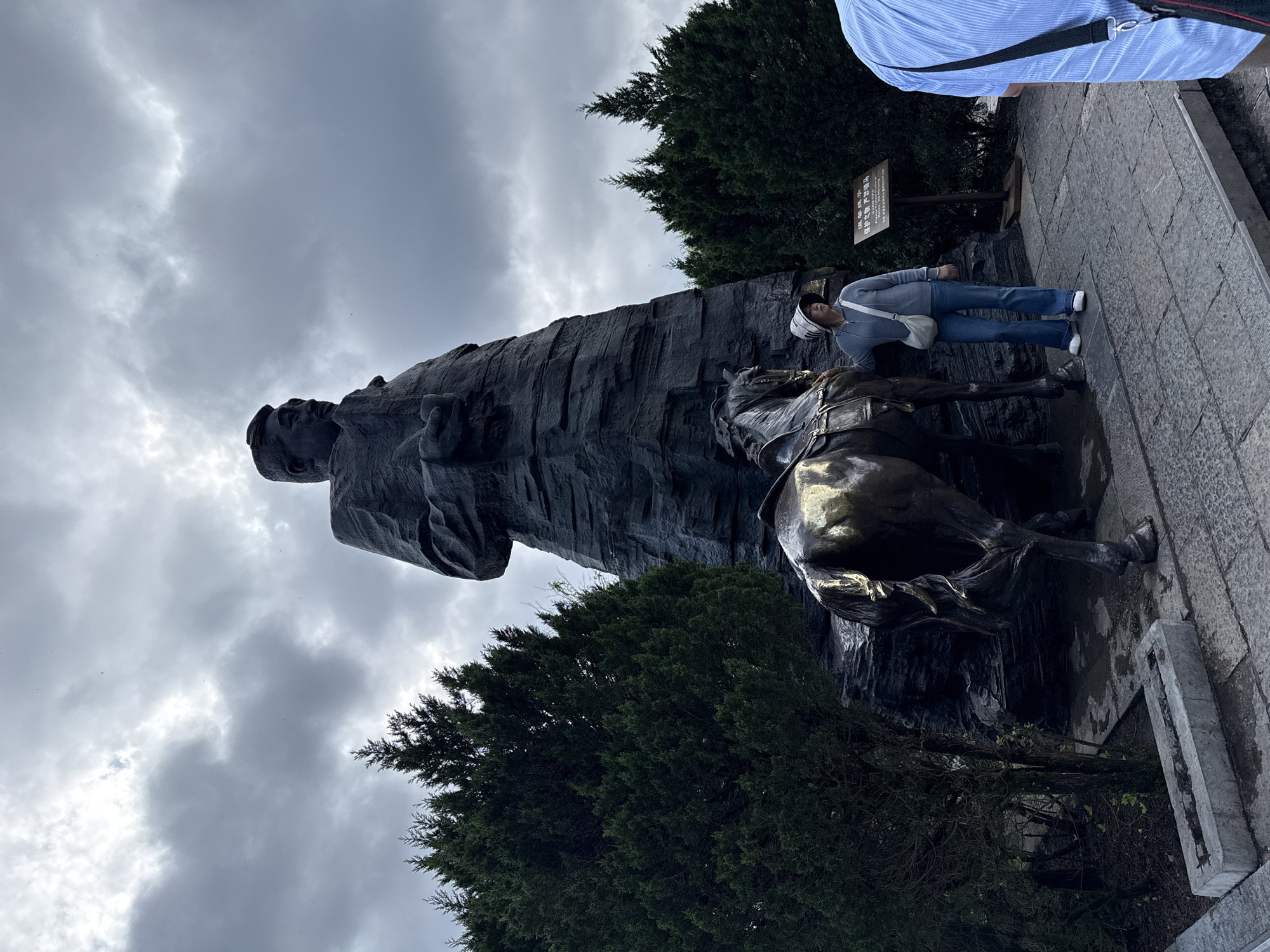 A low-angle, portrait-oriented shot shows a large bronze sculpture of a man on horseback, taken from slightly below eye-level. To the right of the sculpture, a woman wearing a white helmet and gray jacket stands near the horse's leg, leaning slightly against it. A man in a blue shirt is partially visible in the top right corner of the frame. The statue and the woman stand on a paved stone surface. Behind the sculpture, dense green trees fill the lower half of the frame, contrasting with the overcast, cloudy sky above. This photograph was taken in Wulingyuan, China.