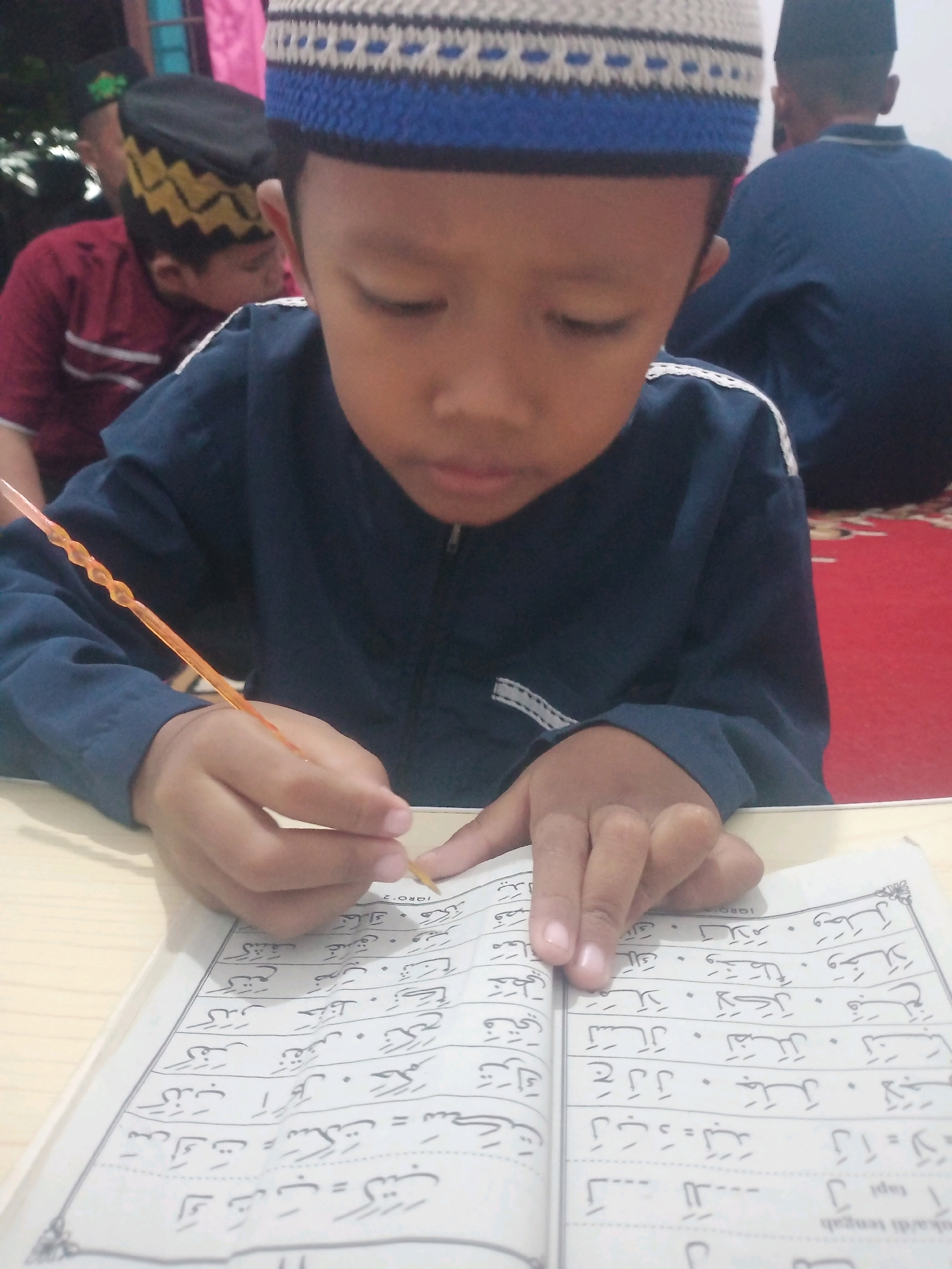 A young boy wearing a blue and white patterned kufi and a dark blue shirt is focused on reading a book with Arabic script, holding an orange stick-like object in his right hand. He appears to be in an indoor setting, possibly a mosque or a classroom, as evidenced by the red carpet visible in the background and other individuals also wearing traditional Islamic attire. The boy's face is partially obscured by his focus on the book, but his brow is furrowed in concentration. The book is open on a light-colored surface, likely a table. The scene captures a moment of religious education.