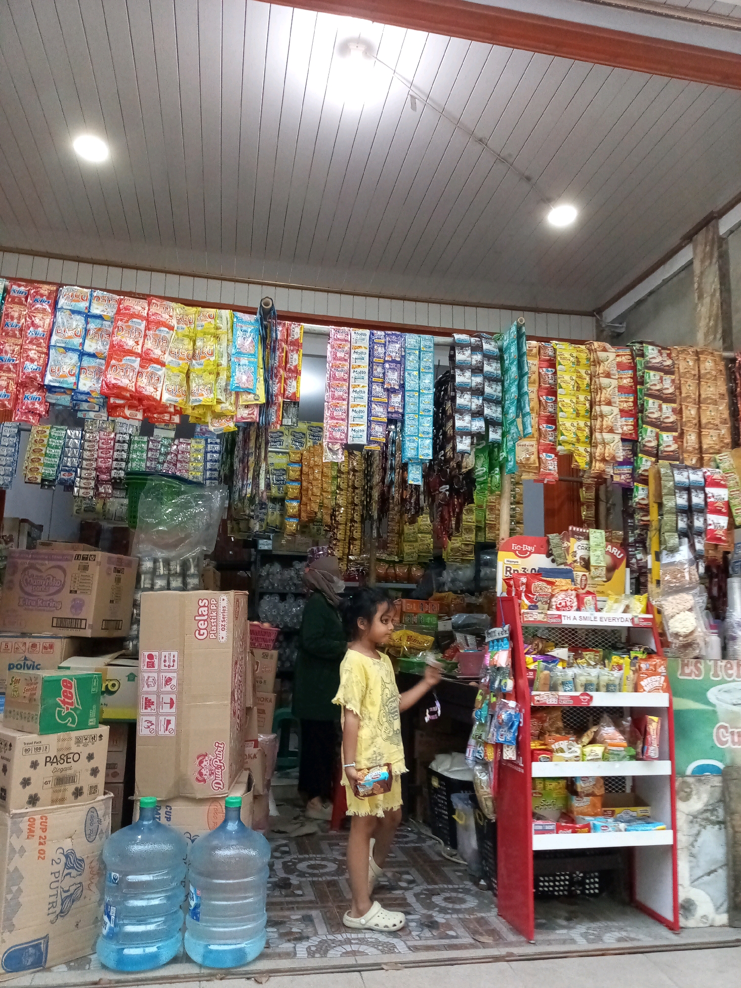 This is a medium shot of a small convenience store in Karangsatria, Indonesia. The store is filled with various packaged goods, including snacks, drinks, and cleaning supplies, displayed on shelves and hung from the ceiling. Two large water jugs are positioned in the foreground, near the entrance. A young girl, wearing a yellow dress and white sandals, stands in front of a red display rack filled with snacks, holding a small package. An adult, wearing a dark hijab and green top, stands behind the girl, partially obscured by merchandise. The store appears to be open, with a bright ceiling and products visible on shelves behind the girl and adult. The floor is tiled with a patterned design.