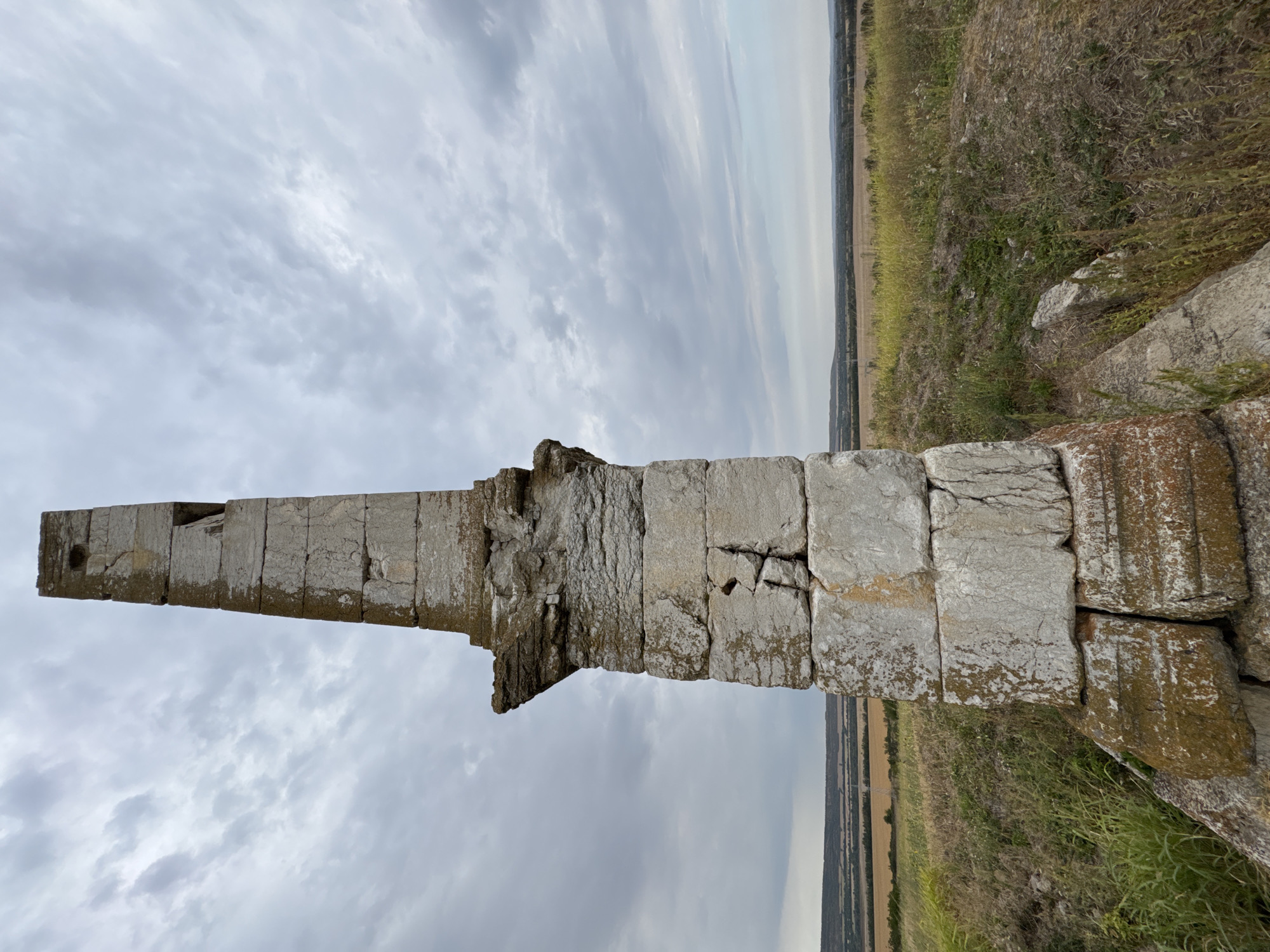 The image shows a weathered stone monument, possibly an obelisk, situated on a grassy knoll. The monument is constructed from large, roughly hewn blocks of light-colored stone, some of which are cracked and show signs of erosion. The top section of the monument tapers and has a rectangular opening. The base of the monument appears to be made of larger, more irregularly shaped stones, some covered in moss or lichen.
The monument is set against a backdrop of a landscape under a cloudy sky. The immediate surroundings are covered in dry grass and scattered green vegetation. In the middle ground, there are fields of dry crops, appearing golden brown, and further in the distance, a line of hills or low mountains can be seen. The horizon is flat and extends across the width of the image. The overall impression is one of an ancient, solitary structure in a rural setting.
This monument is located in the vicinity of Lesicheri, Bulgaria.