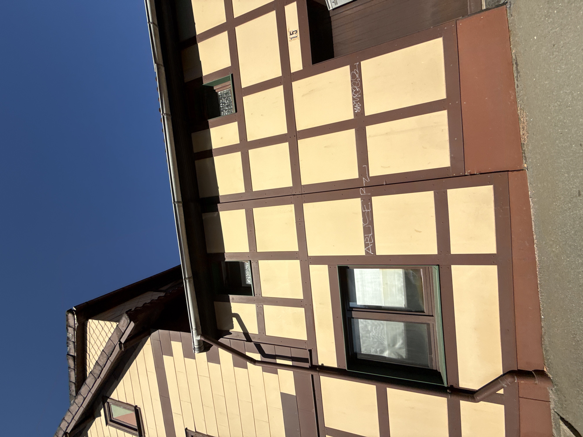 The image captures a close-up, low-angle view of a section of a building in Bad Sachsa, Germany. The building features a distinctive half-timbered architectural style, with a yellow facade and dark brown beams forming a grid pattern. A large downspout runs vertically along the left side of the building.
Several windows are visible on the facade. One on the upper left is small with a triangular pane of glass. Below it is a larger, rectangular window with a dark frame and partially obscured glass, covered by a translucent material. Another small, square window is located higher up on the right section of the facade, with a patterned glass.
Graffiti is present on the yellow sections of the facade. On the lower left, white graffiti reads "ABUSE P.Z." To the right of this, more white graffiti is visible, appearing as a stylized tag or signature. Near the top right of the visible facade, the number "15" is painted in black, likely a house number.
The roofline is visible in the upper left of the image, with dark shingles and a prominent eave. The sky above is a clear, deep blue. The ground in the foreground is a greyish, textured surface, possibly a sidewalk or pavement. The overall impression is of a sunny day, highlighting the colors and textures of the building.