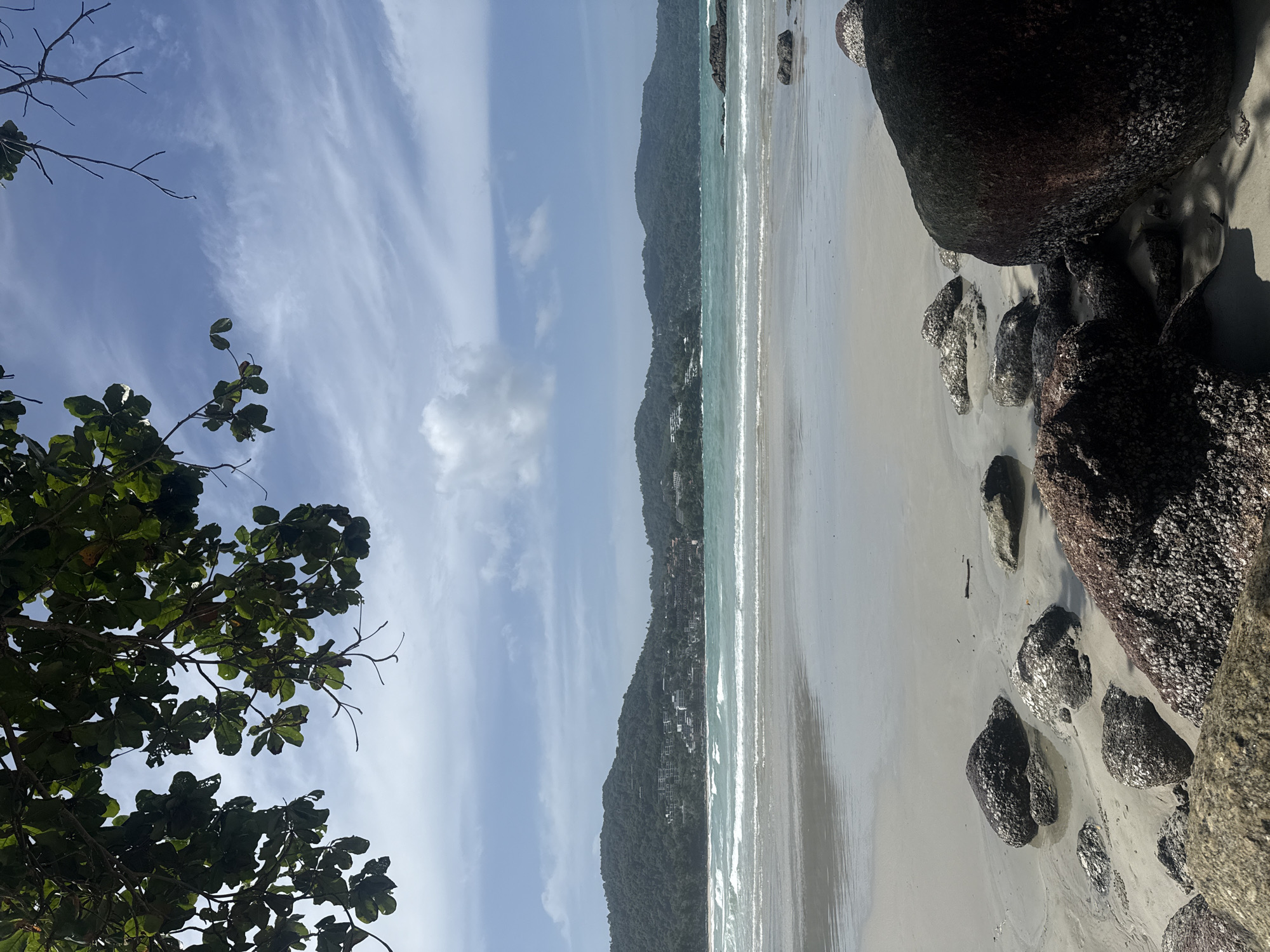 An overcast, bright day at a beach in Karon, Thailand, shows a view across a sandy shore with scattered rocks, some encrusted with barnacles. The ocean exhibits gentle waves and a turquoise hue. In the distance, a range of green, tree-covered hills dotted with buildings creates the coastline. Overhead, the branches of a leafy tree frame the top of the image, with its dark green leaves contrasting against a pale blue sky streaked with thin, wispy white clouds.