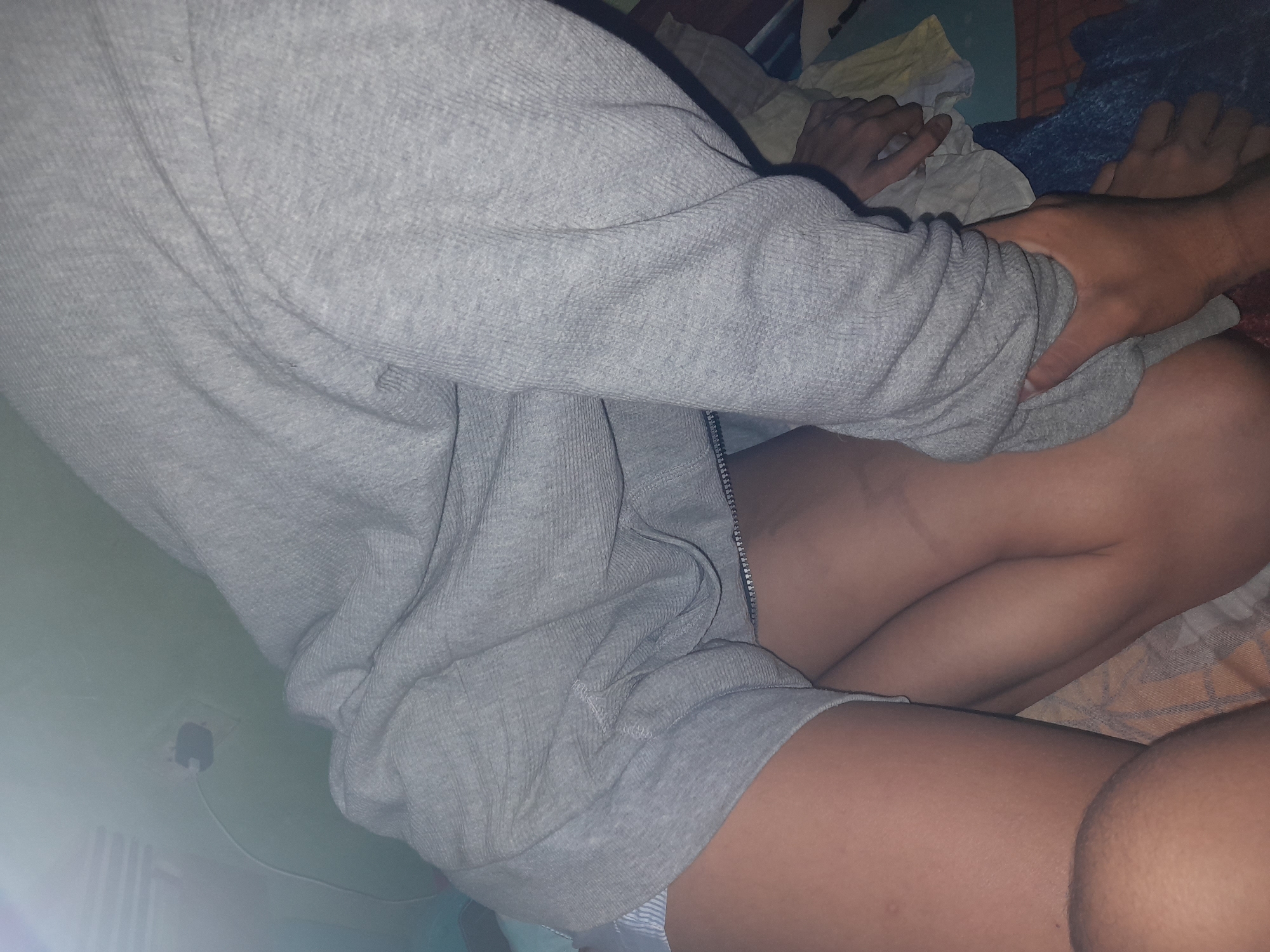 In an indoor environment, possibly a bedroom, two individuals are partially visible. One person, wearing a grey hooded sweatshirt and white striped shorts, is lying down. Their legs are exposed from the upper thigh down. Another person's hands are visible interacting with the first person's sweatshirt and leg. The background shows a light green wall with a black electrical outlet and cord. The lighting is dim, suggesting evening or nighttime. The scene does not provide any specific indicators to determine the location within Santa Ana, Venezuela.