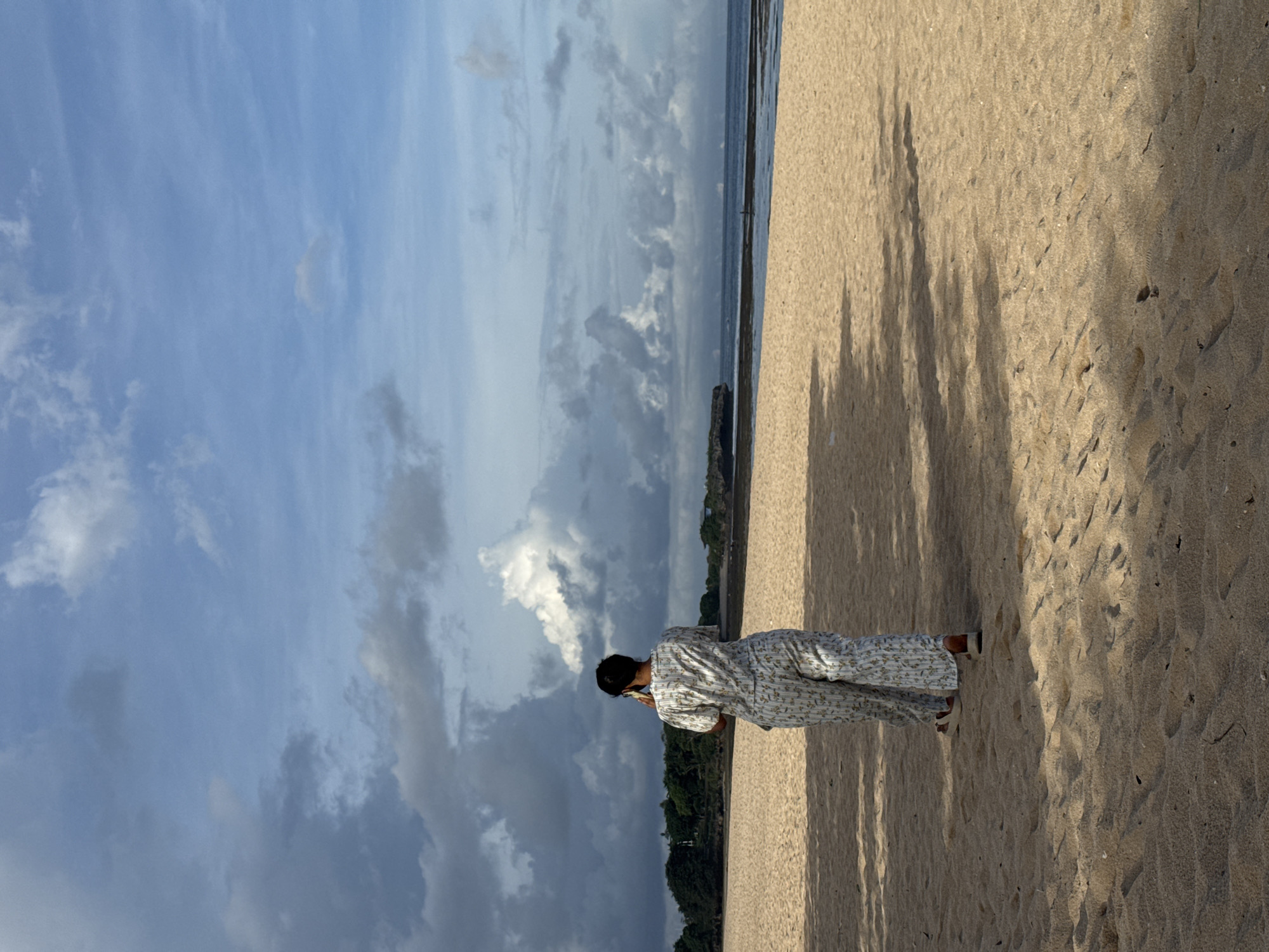 A woman stands on a sandy beach under a partly cloudy sky. She is wearing a light-colored patterned outfit and appears to be using a mobile phone. The beach has tree shadows, and the ocean is visible in the background. The location is Kuta Selatan, Indonesia.